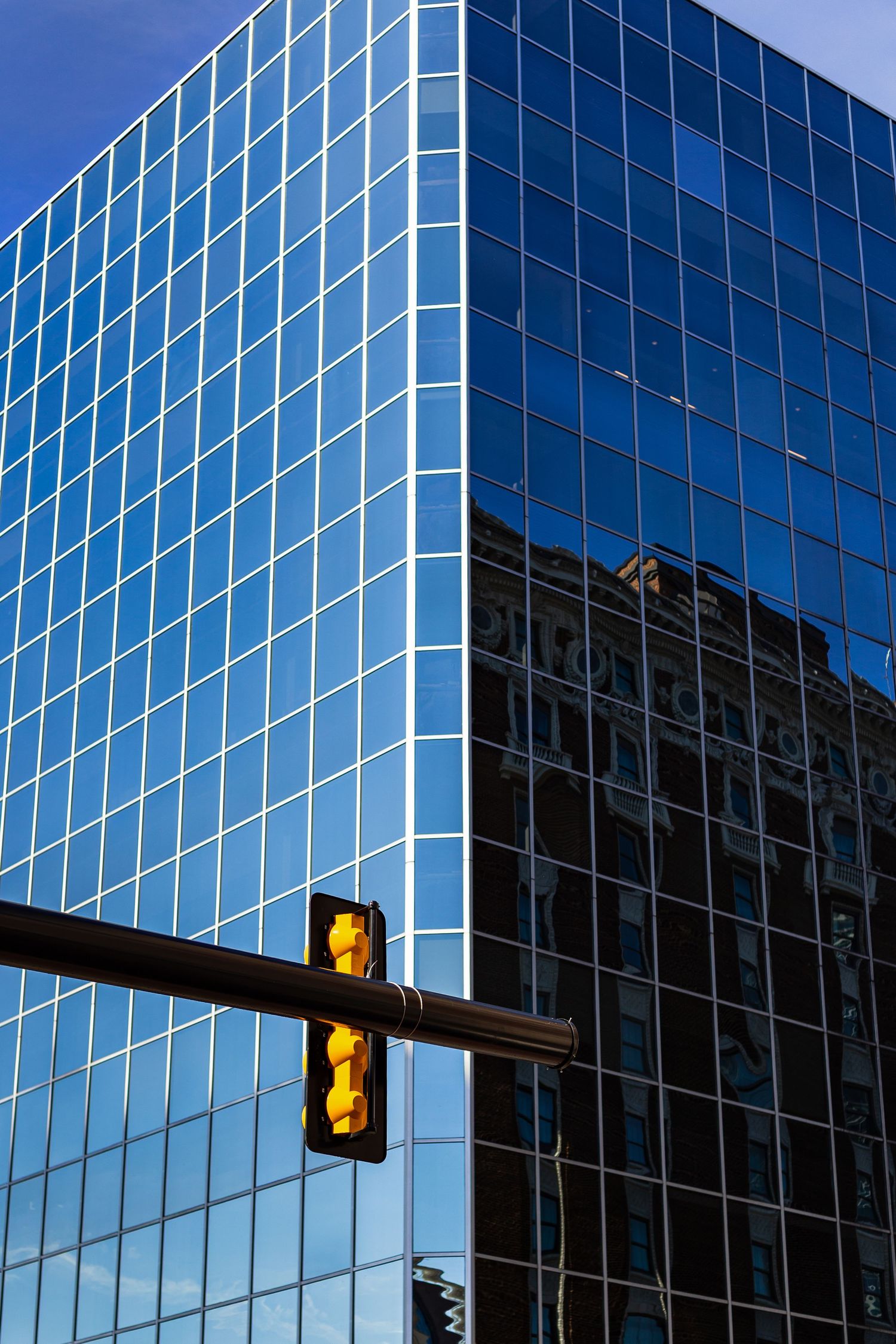 Modern glass skyscraper reflecting an ornate historic building, with a yellow traffic signal in the foreground against a blue sky.