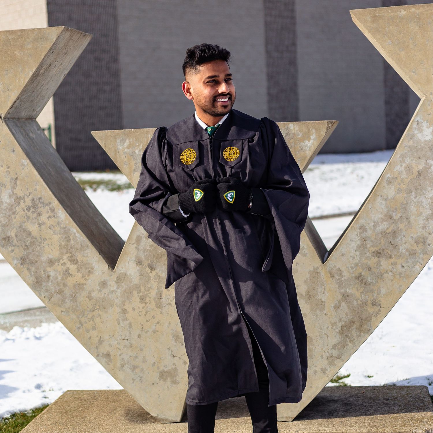 Graduate in black gown with patches posing by W sculpture.