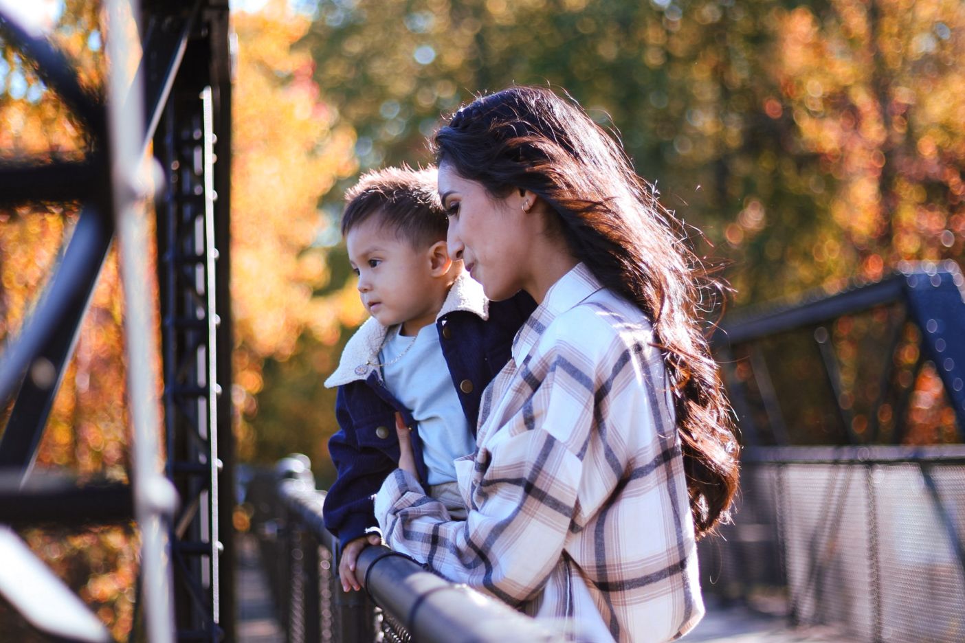 A young woman holds a toddler while leaning on a bridge railing surrounded by autumn foliage.