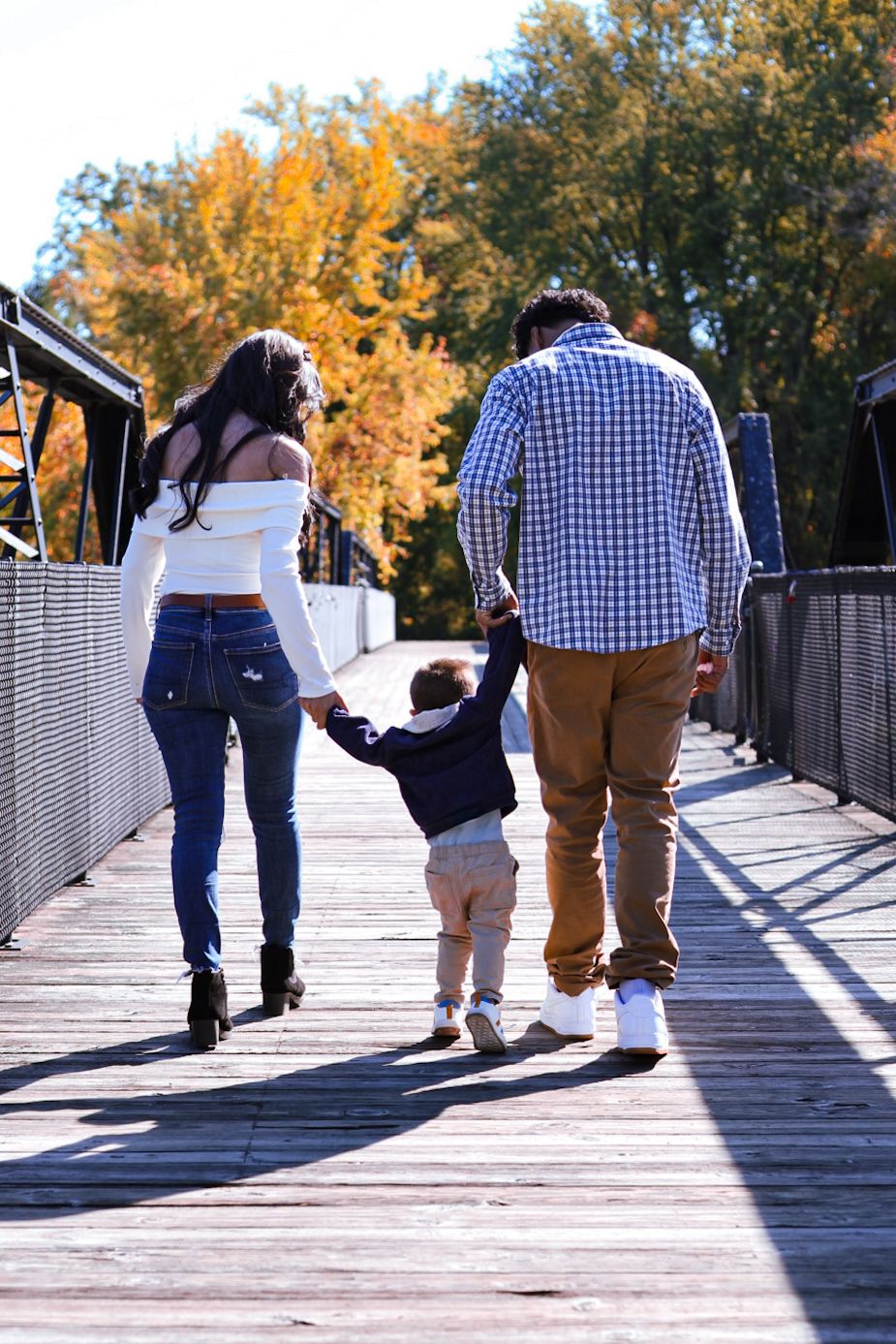 African American family walking hand-in-hand on a wooden bridge surrounded by colorful autumn foliage.