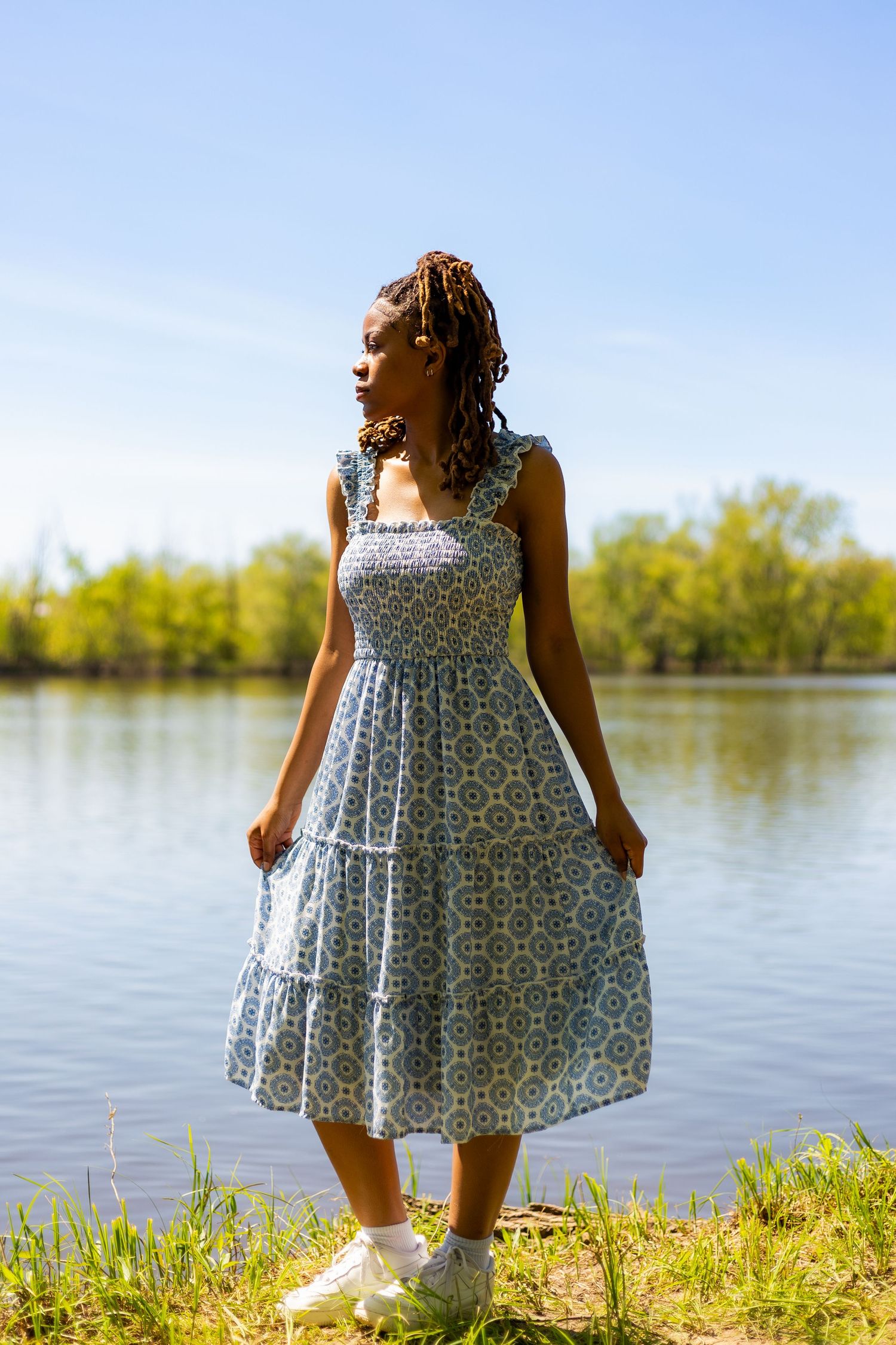Young Black woman in blue floral midi dress standing by a calm lake on a sunny spring day.