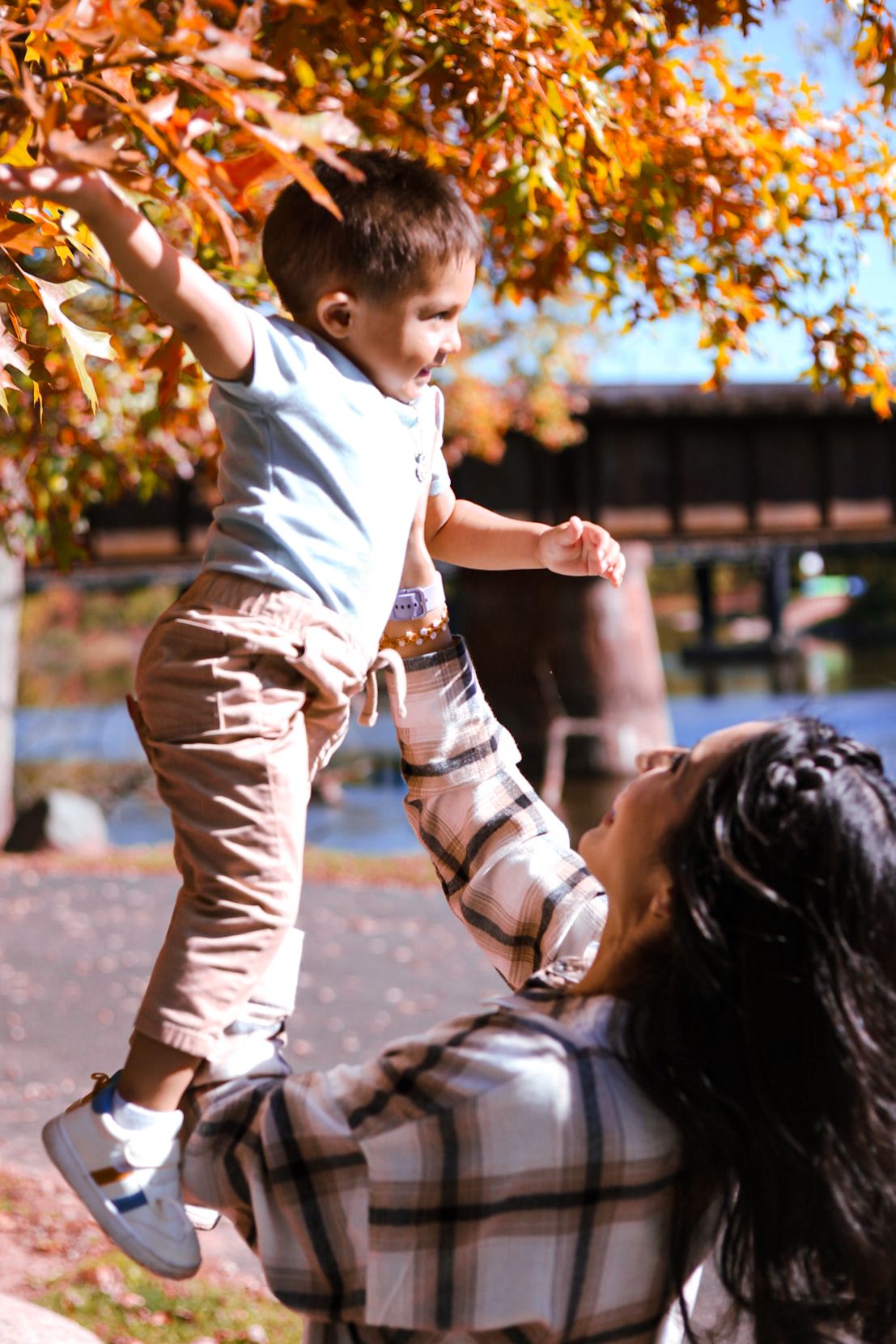 A joyful mother lifts her laughing toddler son toward colorful orange autumn leaves by a lake.