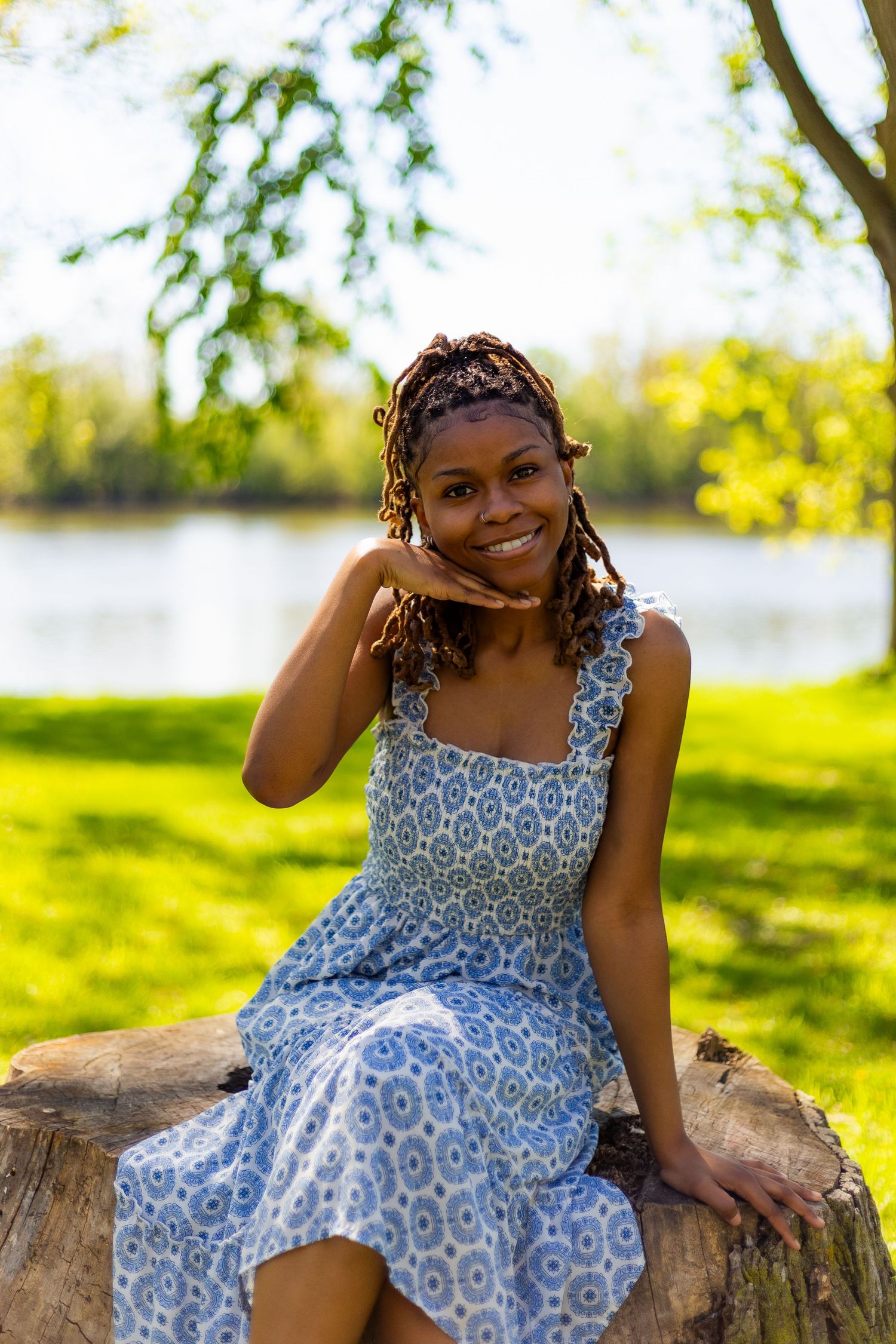 Happy young Black woman in blue sundress smiles while sitting on tree stump near lake.