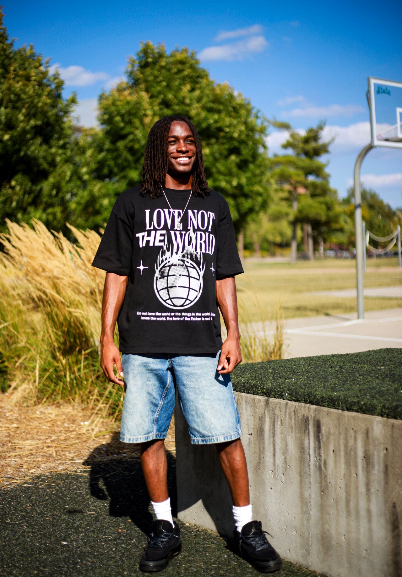 Smiling man in black 'Love Not The World' tee and denim shorts standing by outdoor basketball court.