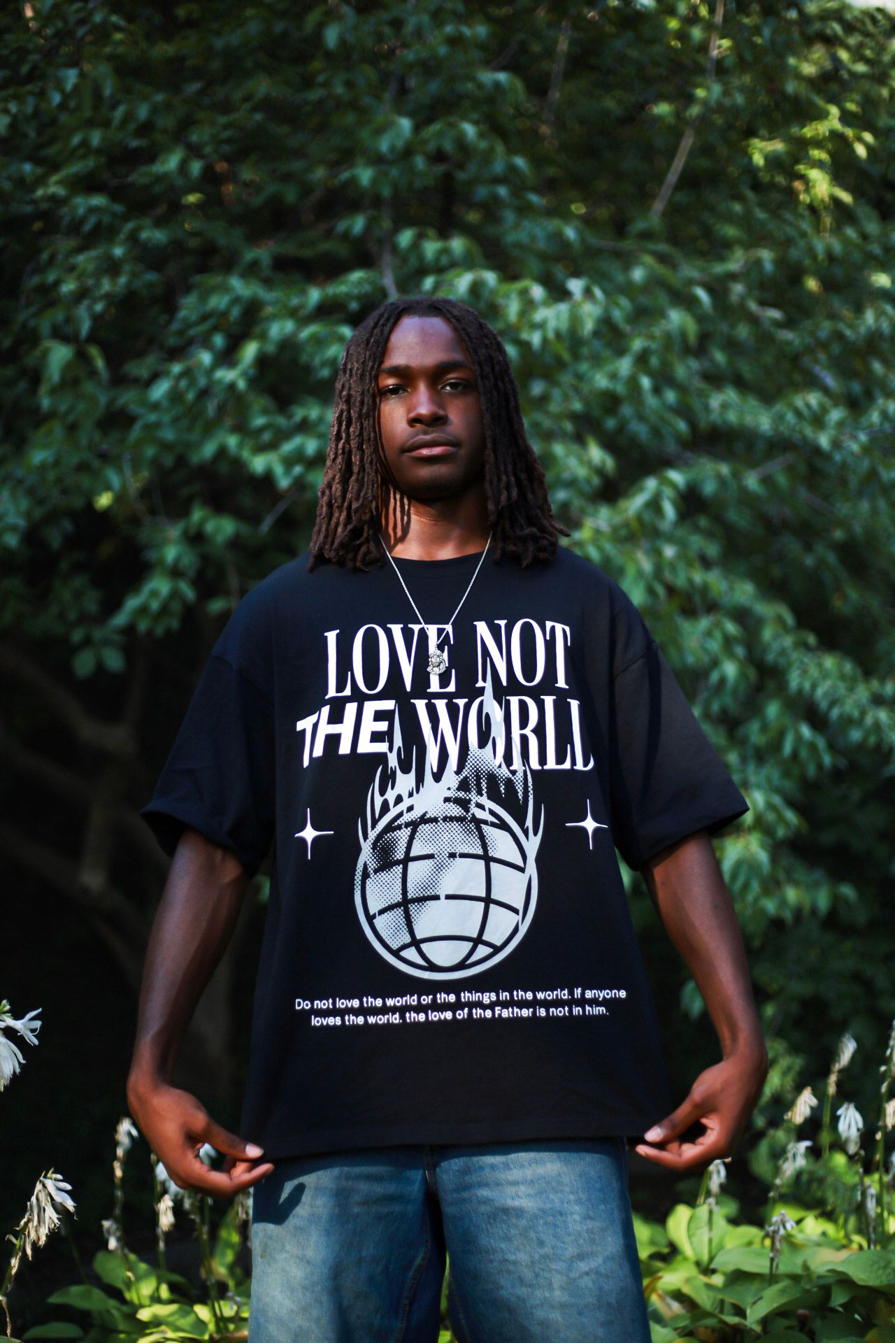Man modeling black Love Not The World oversized graphic tee surrounded by green foliage.