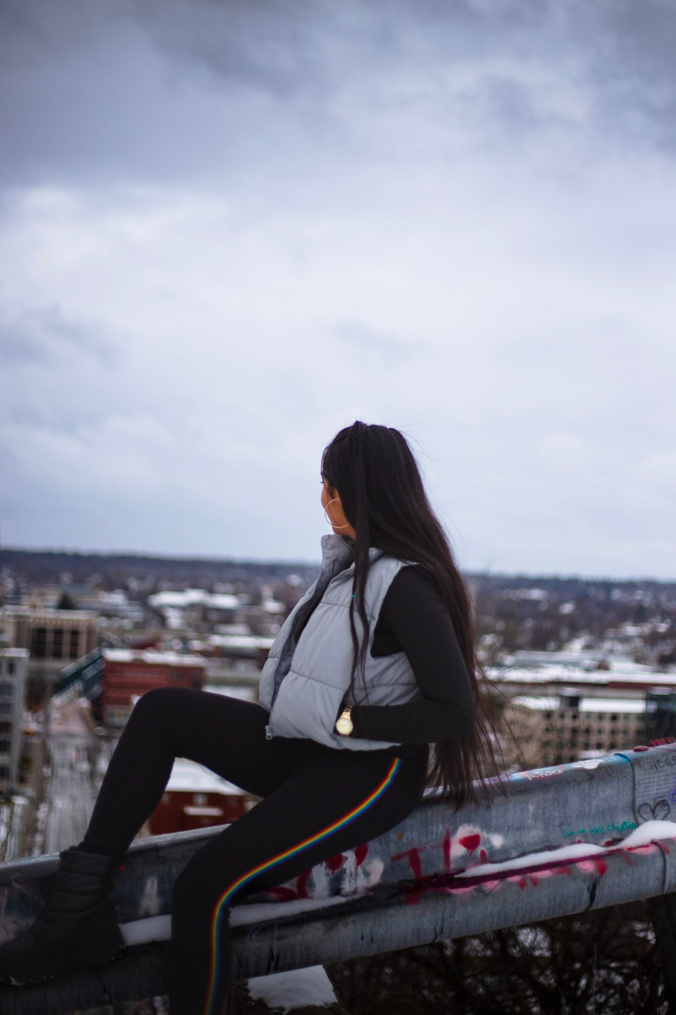 Woman in white vest sitting on railing with panoramic snowy city view behind her.