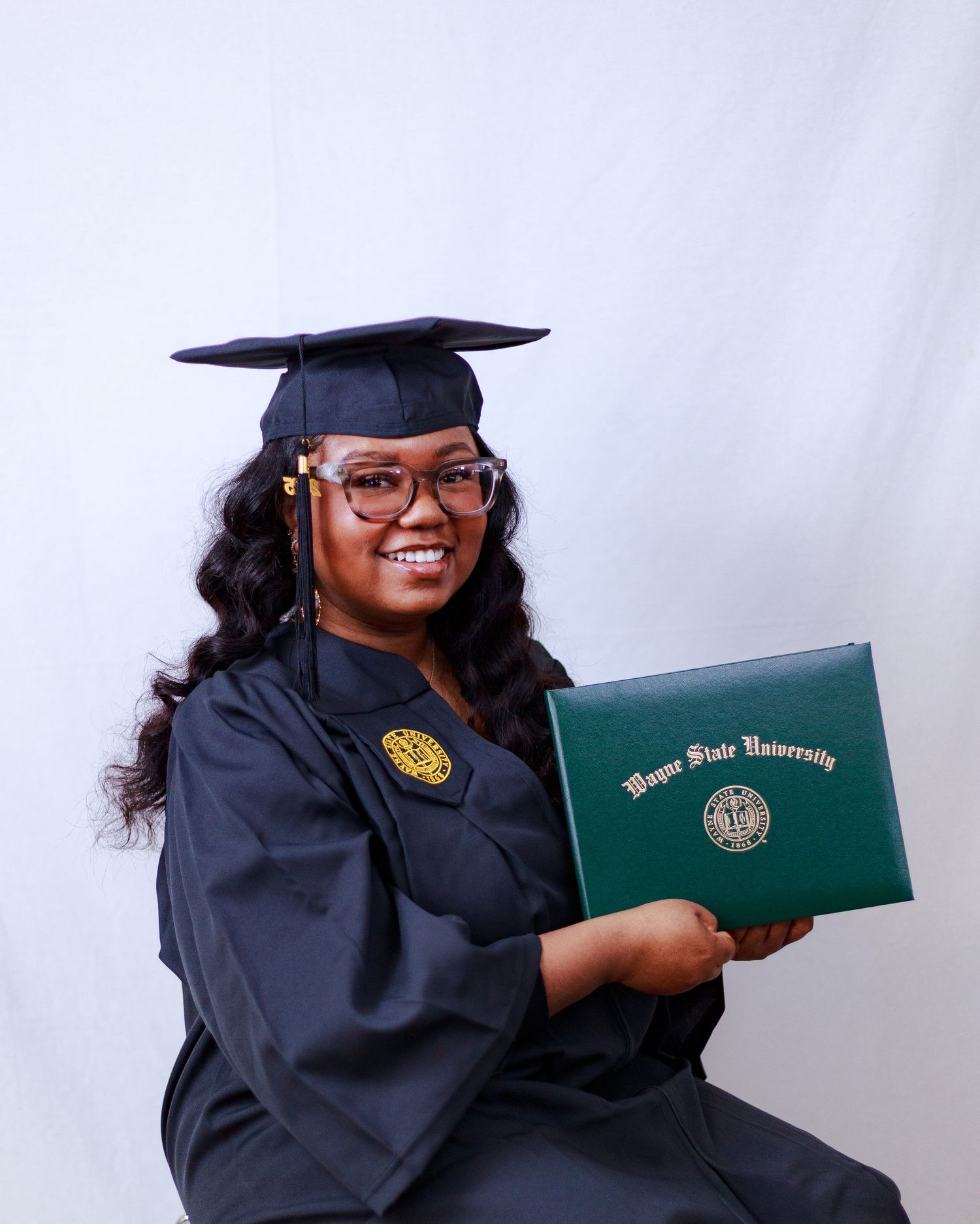 Smiling female graduate holding Wayne State University diploma while seated in cap and gown.