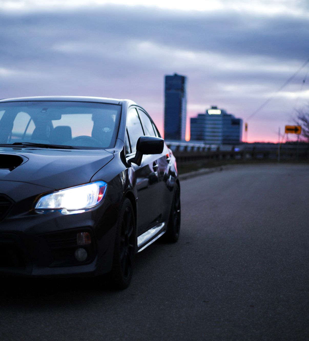 Black Subaru WRX STI parked at dusk with city skyline and dramatic purple clouds in background.