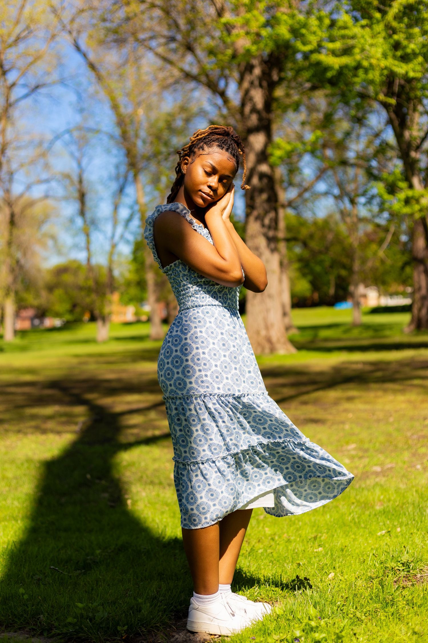 Young Black woman in blue floral midi dress poses gracefully in sunny park with lush green trees.