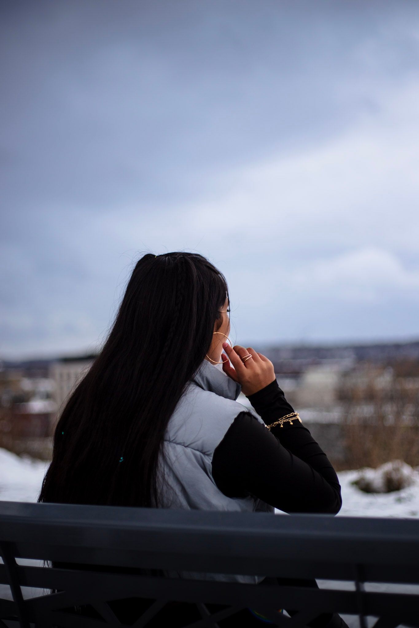 Woman with long black hair sitting on bench overlooking snowy winter cityscape on cloudy day.