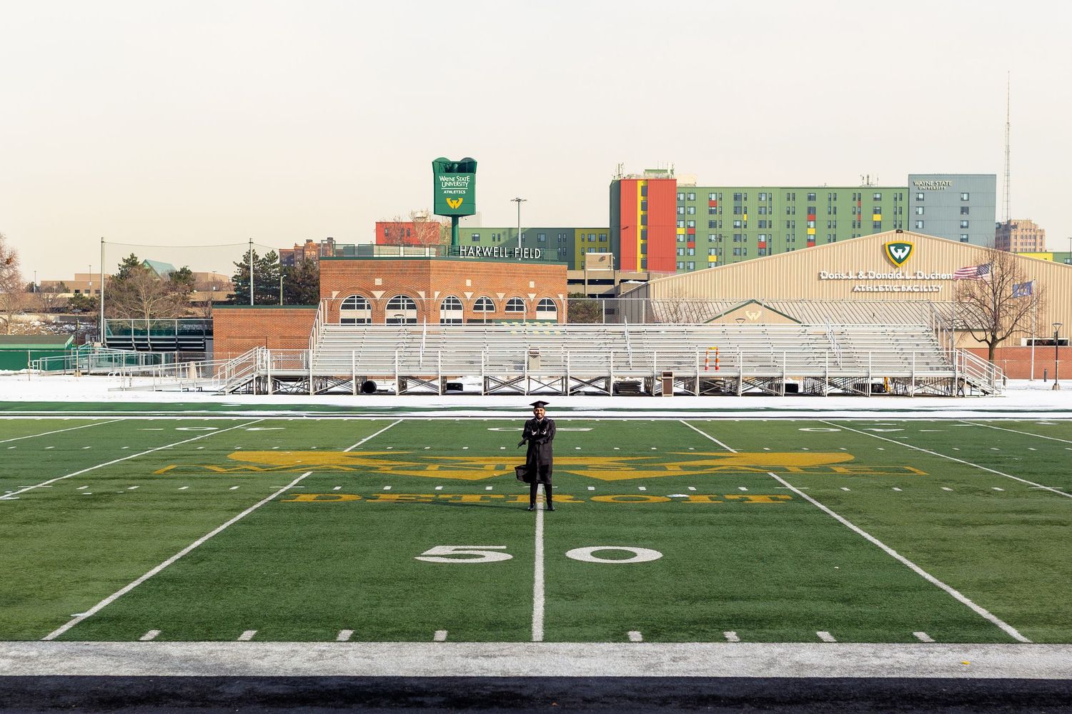 Graduate standing alone on a snowy football field at dusk.