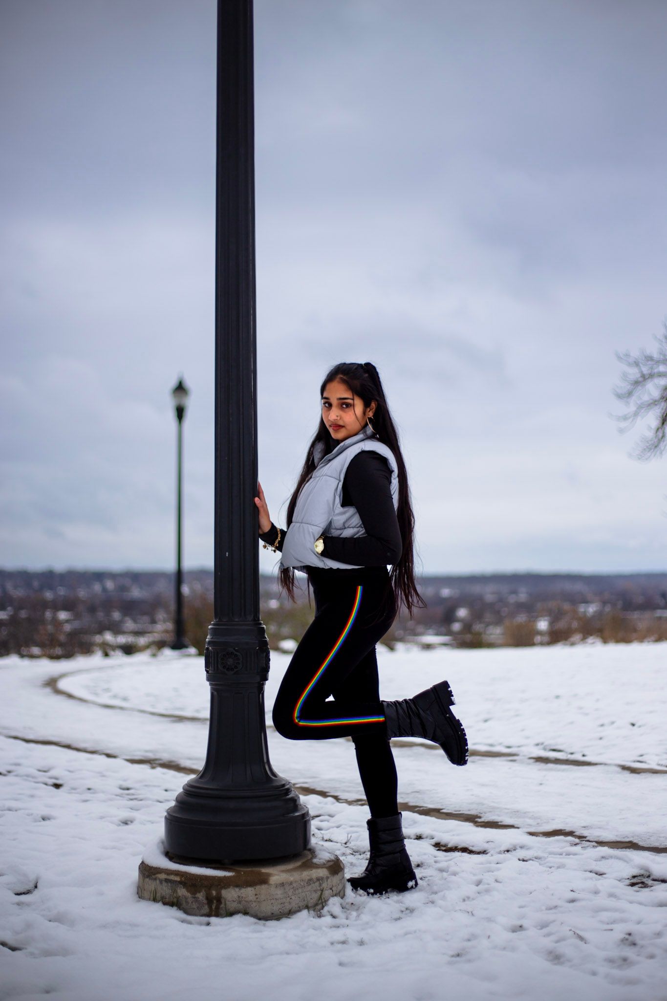 Young woman in silver vest and black leggings poses by a lamppost in a snowy winter park.