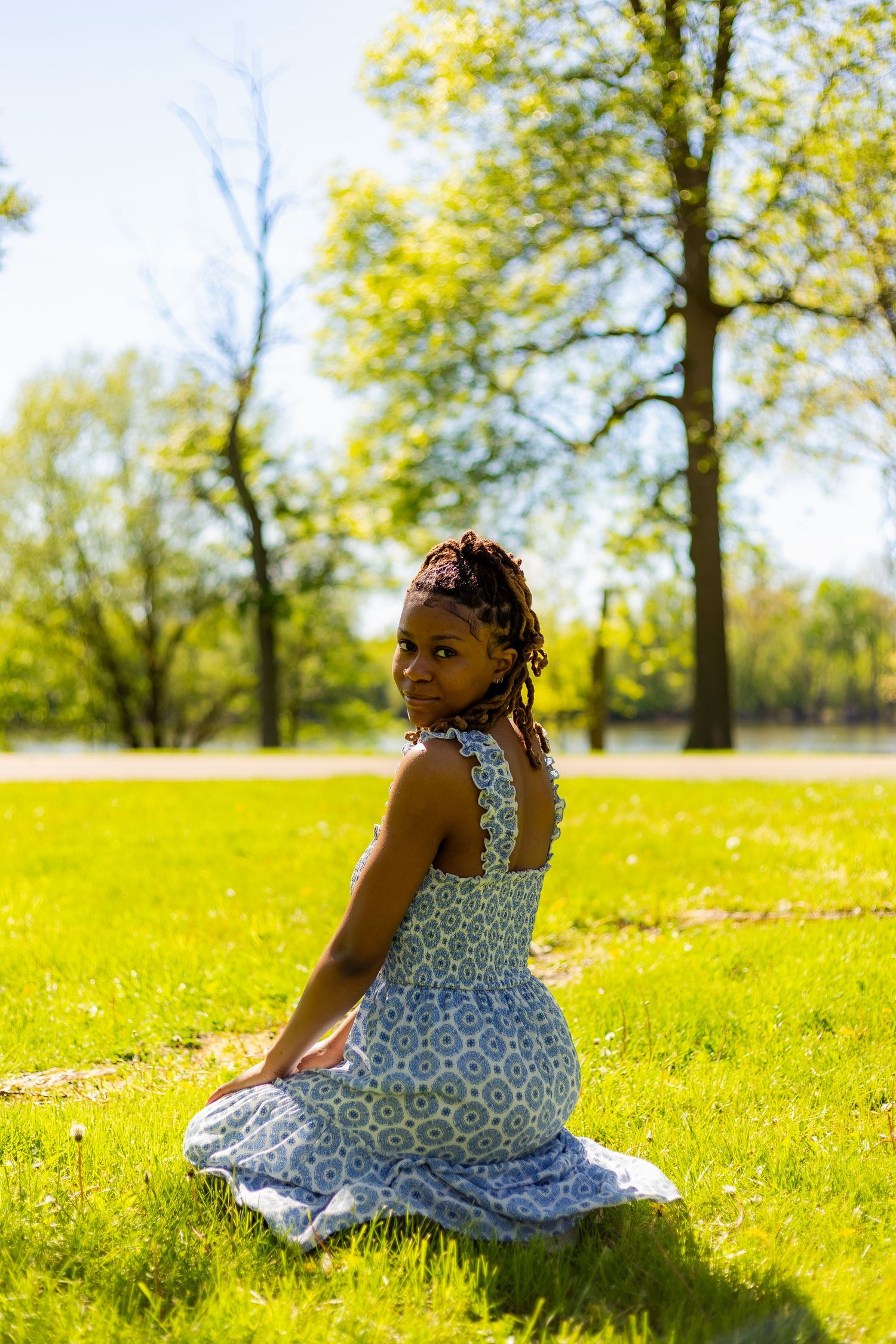 Young Black woman in blue floral maxi dress sitting on lush green grass in sunny park with trees in background.