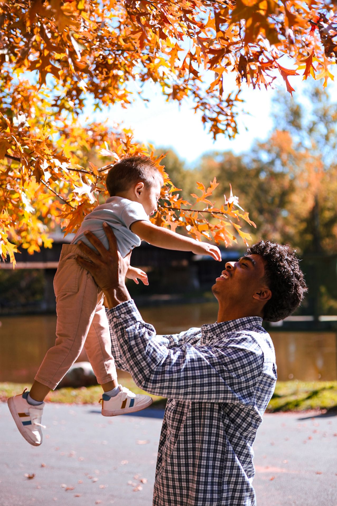 A smiling father lifts his happy toddler son high in the air under golden autumn foliage by a lake.