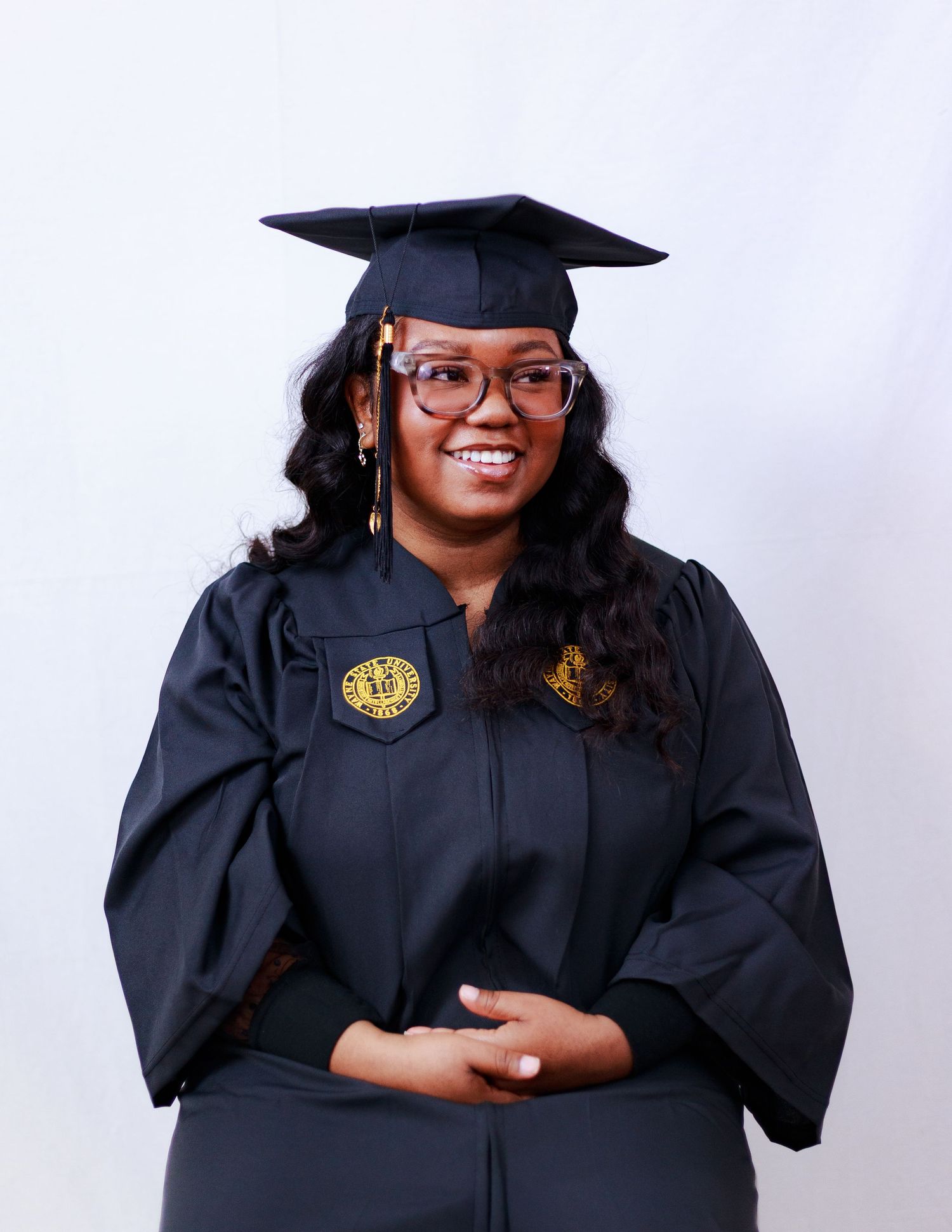 Smiling graduate in black cap and gown with gold emblem posing for portrait against white background.