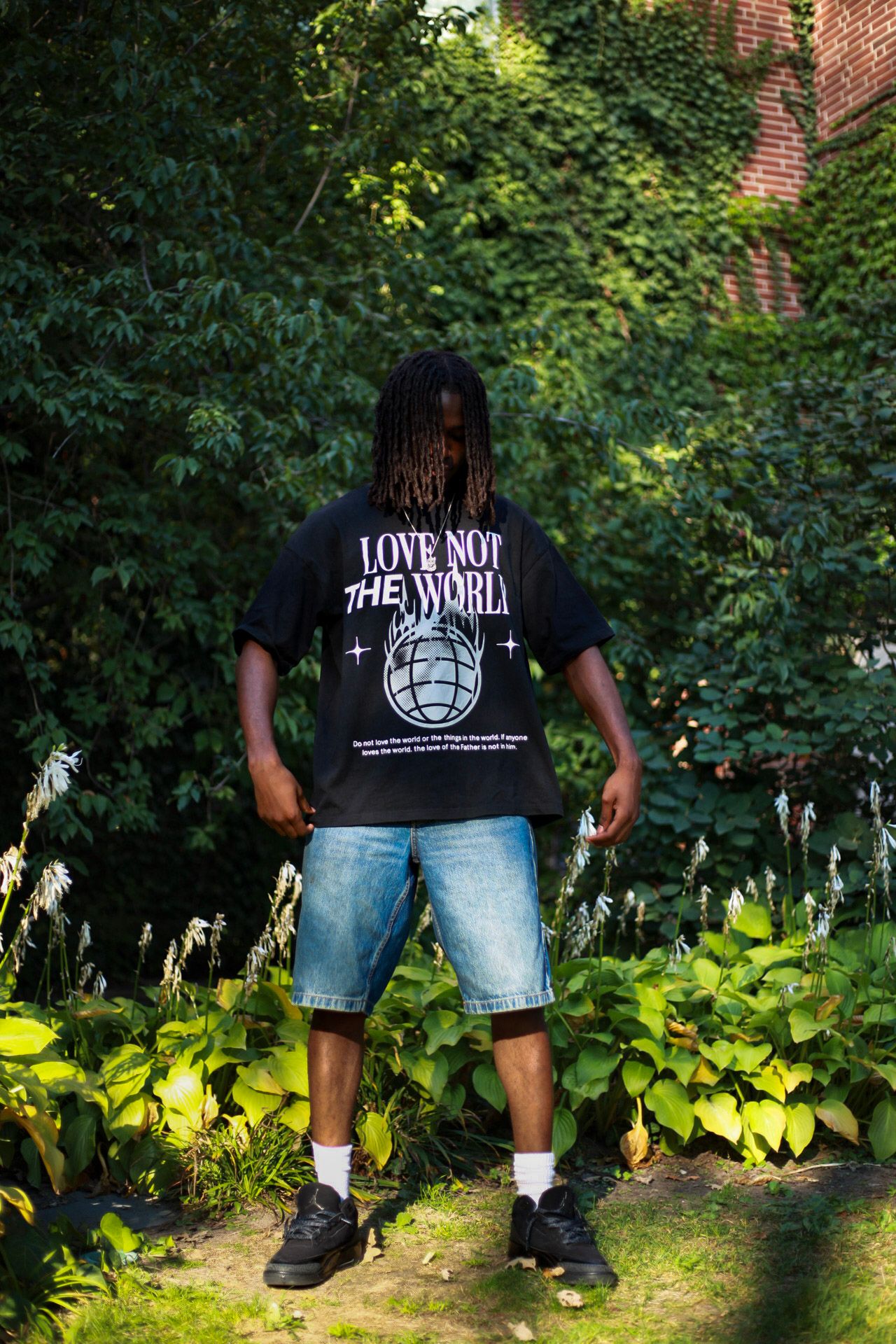 Man showing back of black 'Love Not The World' graphic tee wearing denim shorts in garden setting.
