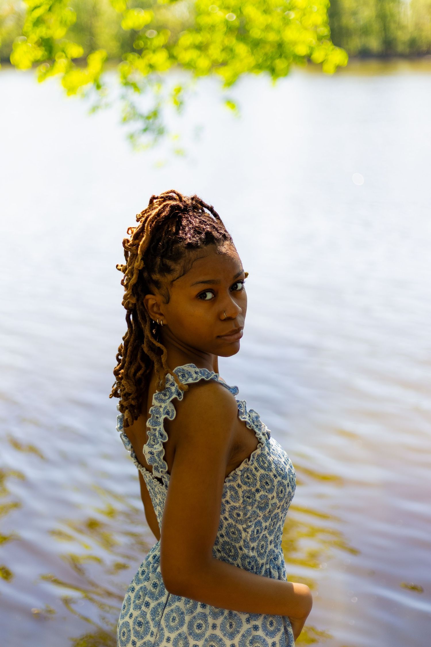 Young Black woman in blue floral dress glancing back over shoulder while standing near a sunlit lake.