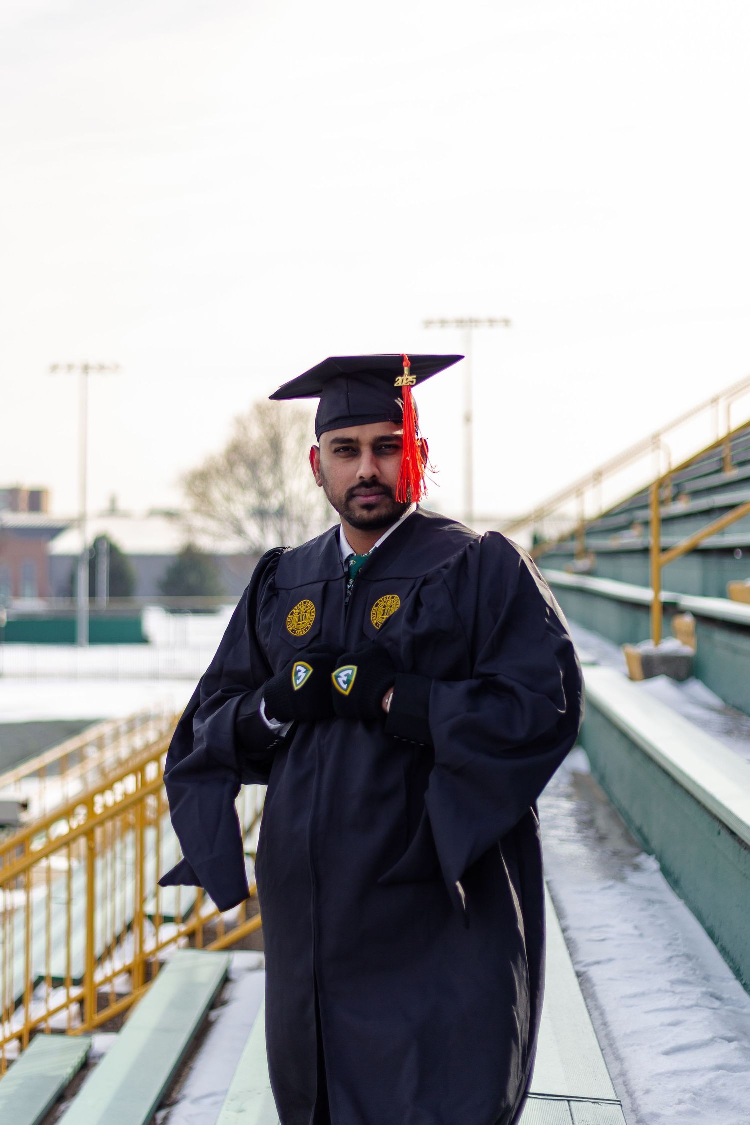 Graduate in black Wayne State University cap and gown poses in snowy stadium bleachers.