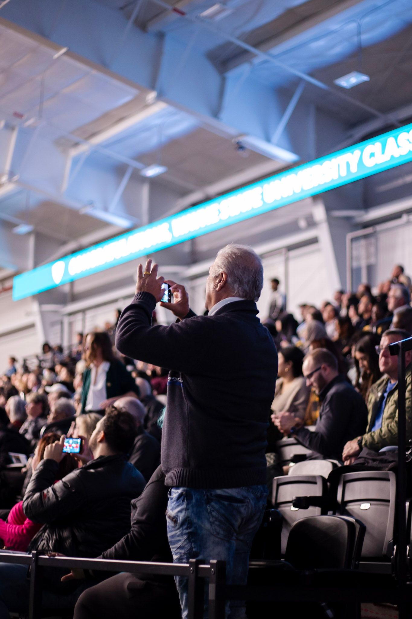 Man standing in crowded arena bleachers photographing a Wayne State University graduation ceremony on his smartphone.