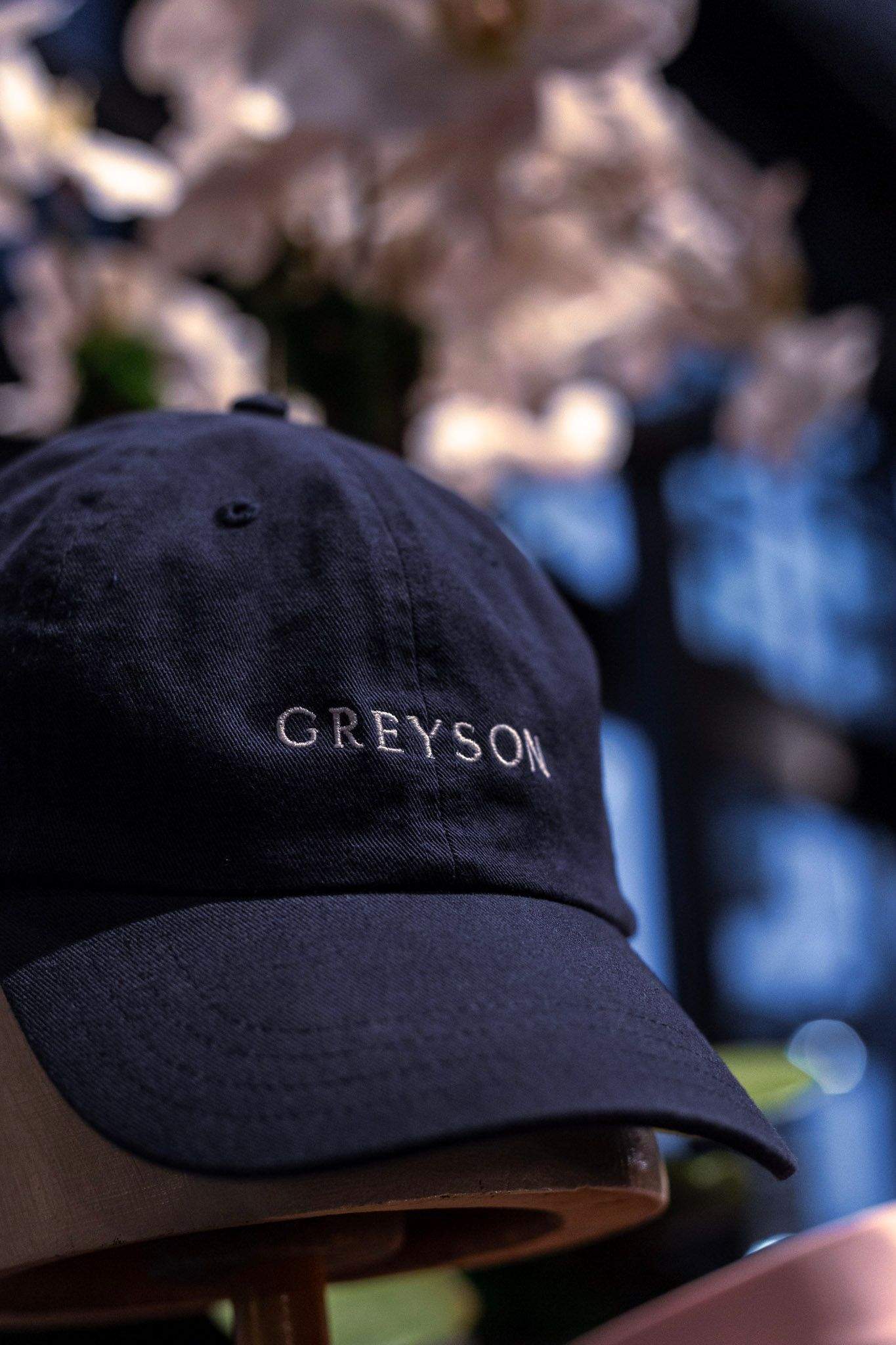 Navy blue Greyson branded dad hat with embroidered logo displayed in front of soft white floral background.
