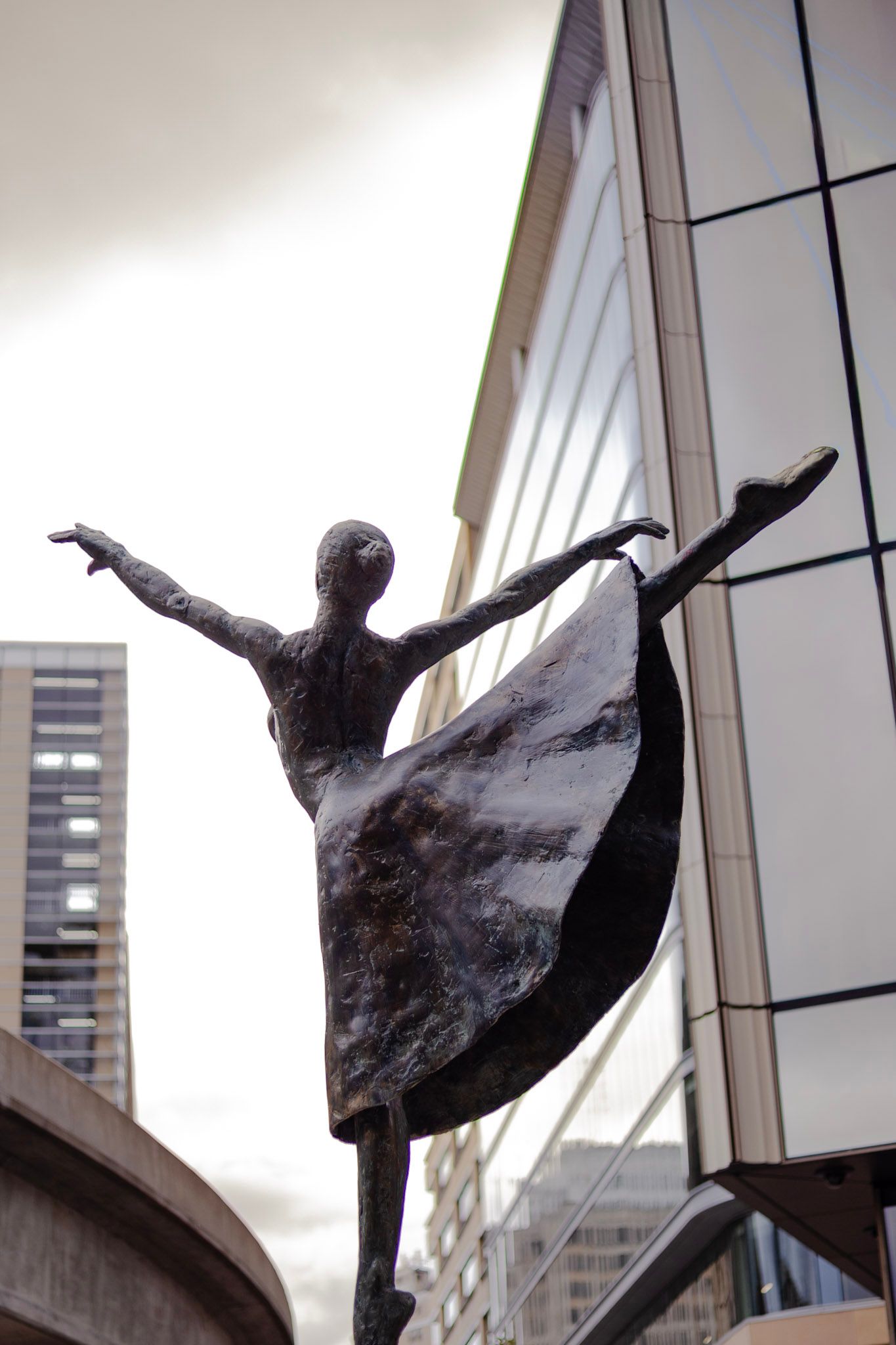Bronze ballerina dancer sculpture in arabesque pose with modern glass building and city skyline backdrop.