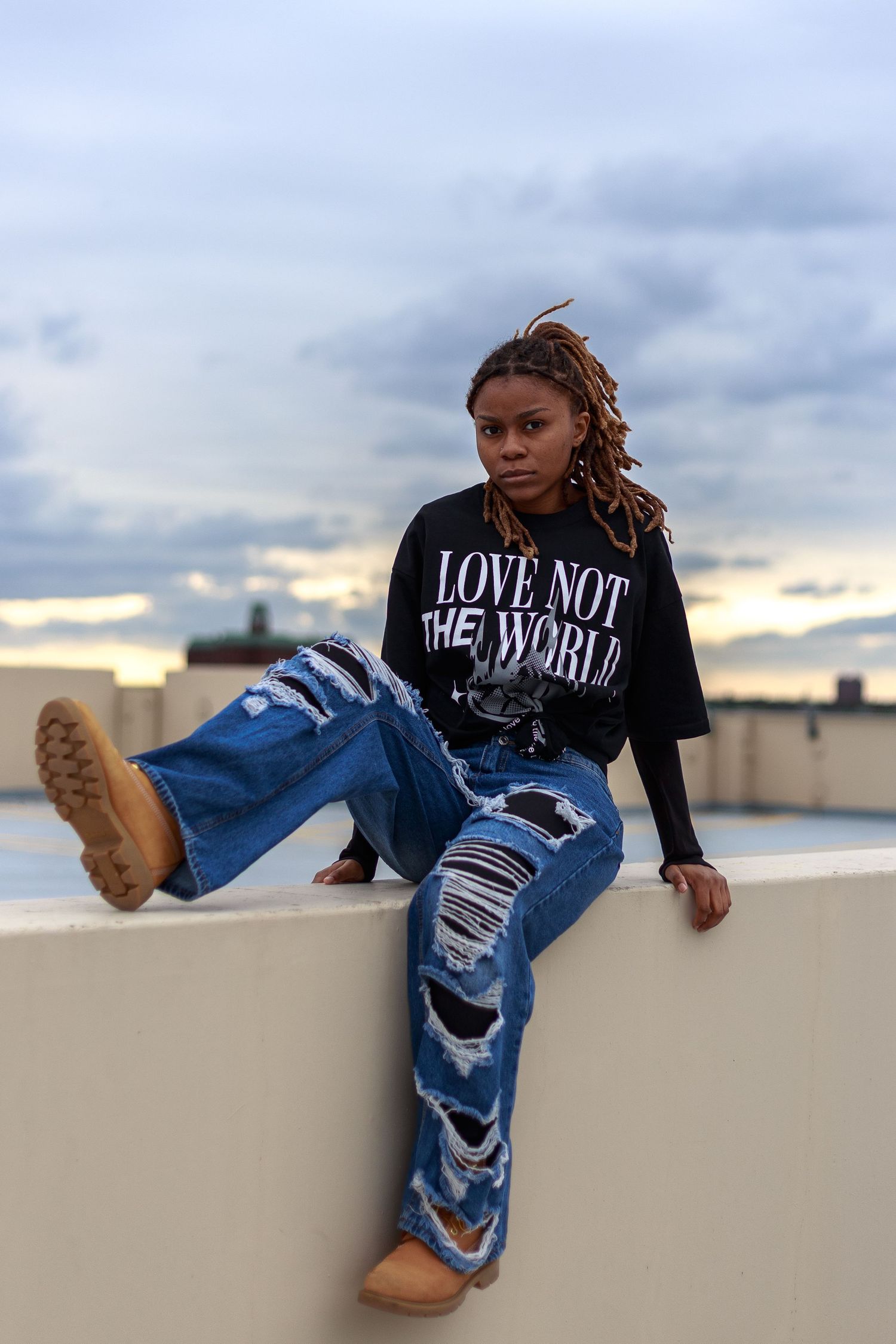 Woman in ripped wide-leg jeans and 'Love Not The World' tee sits on rooftop ledge at dusk wearing tan boots.