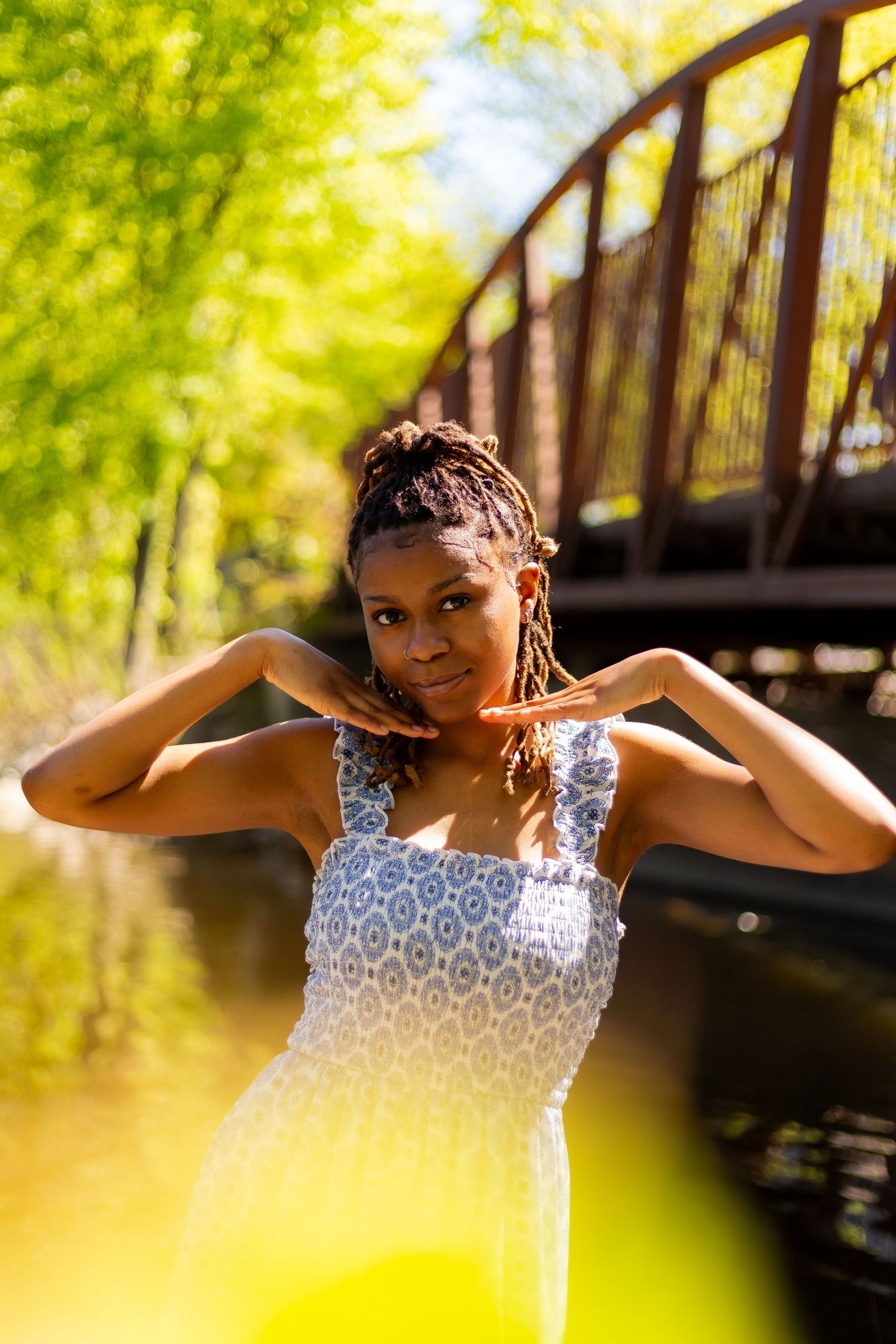 Young Black woman in blue sundress poses confidently near rustic bridge in sun-dappled park.