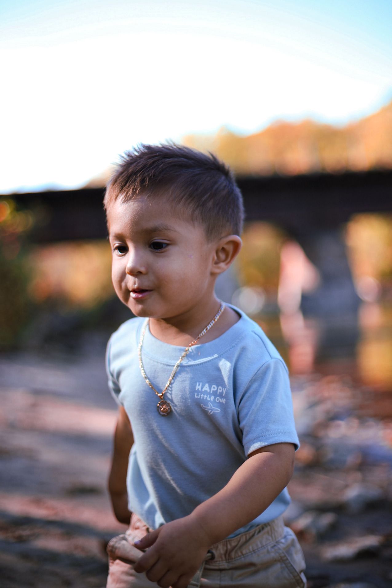 A young boy wearing a white Happy Little One shirt and gold necklace stands outdoors near a bridge.
