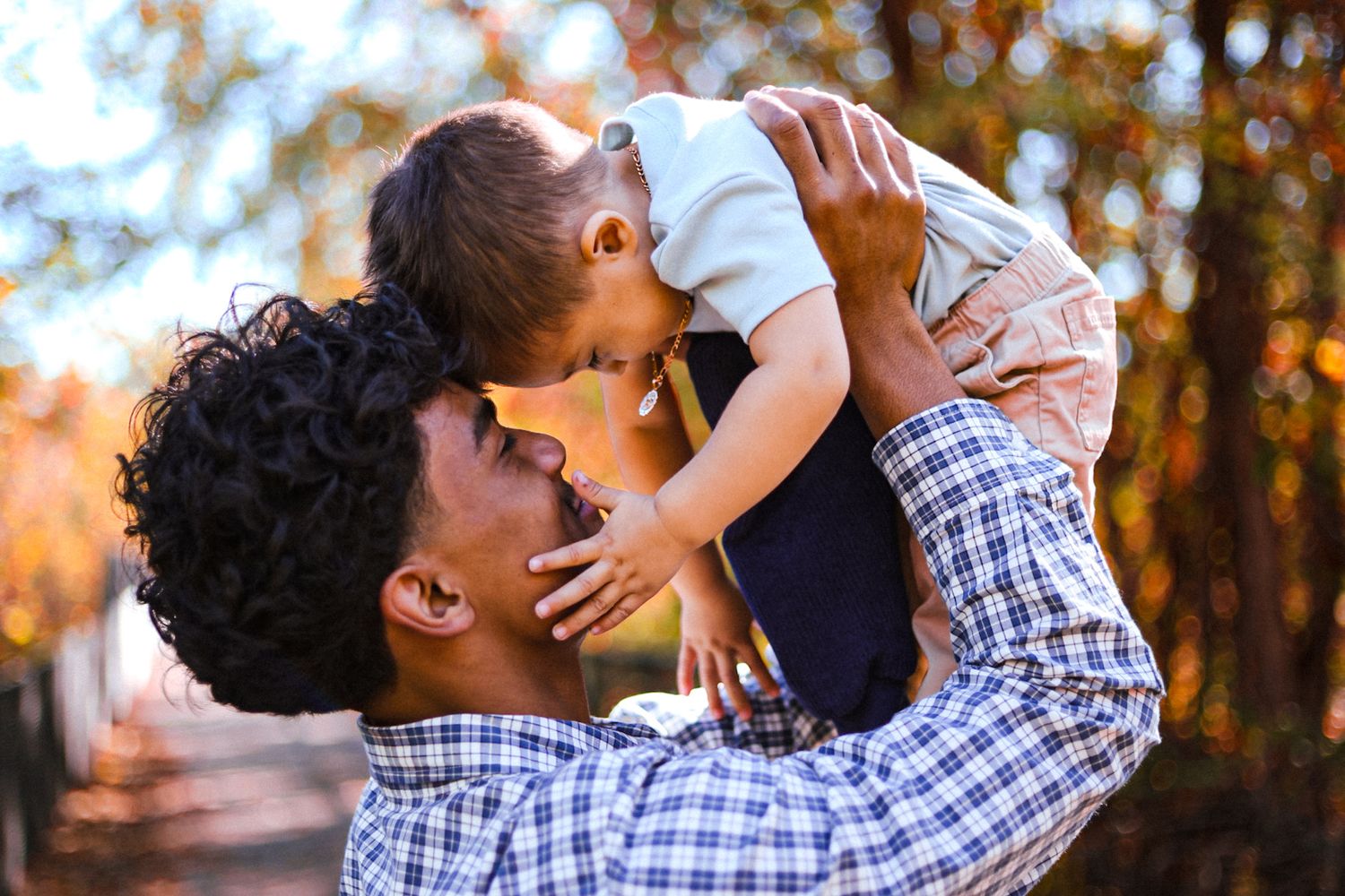 Father lovingly lifting his smiling toddler son up in a colorful autumn outdoor setting.