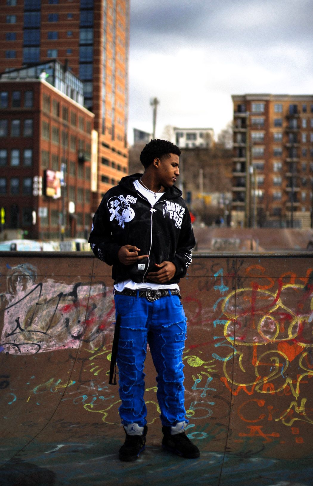 Young man in blue cargo pants and black graphic bomber jacket posing at urban skate park.