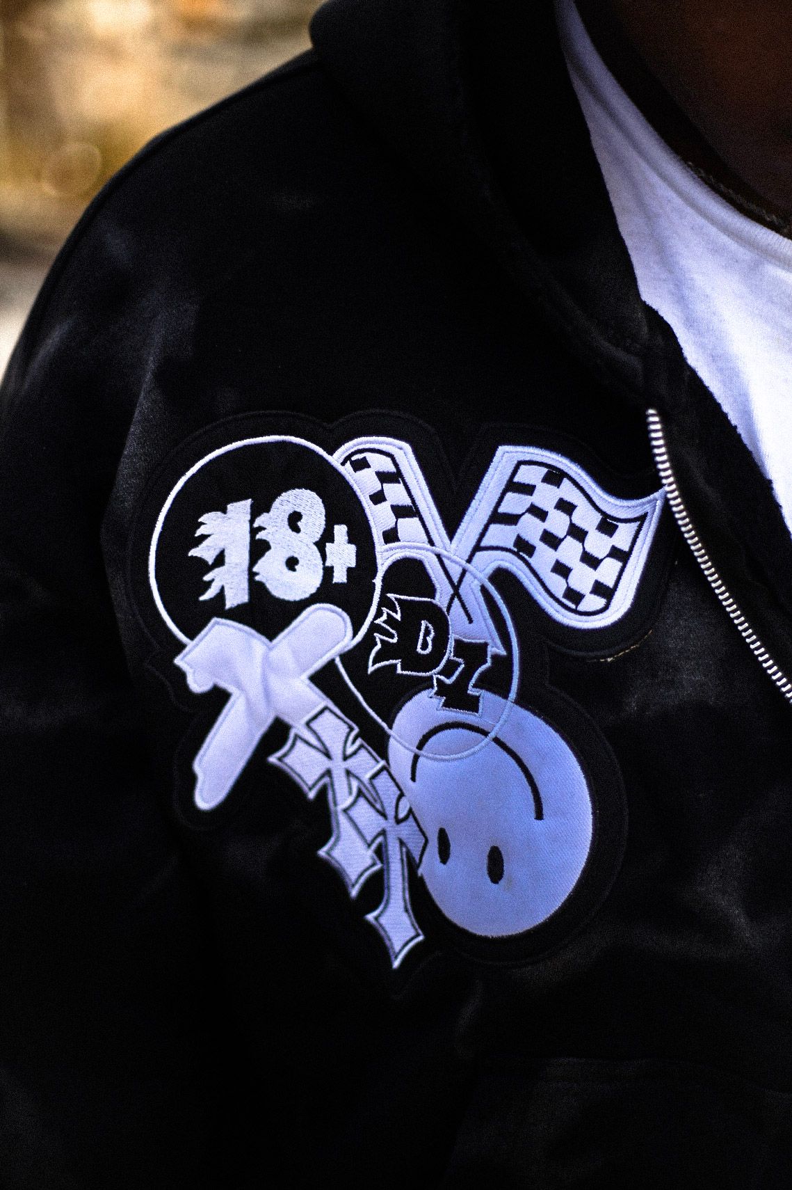 Close-up of black varsity jacket with 18+ racing flags and smiley face graphic patch design.