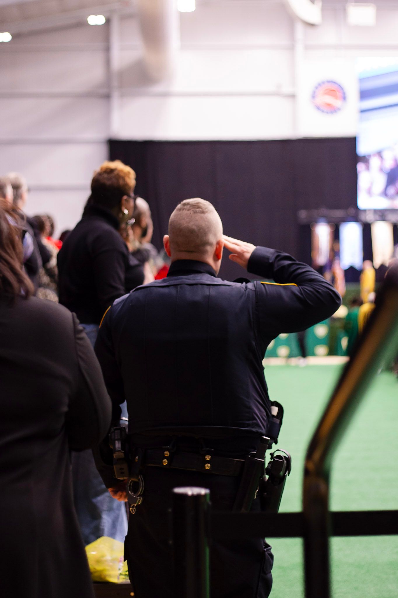 A police officer salutes at an indoor event while attendees gather in the background.