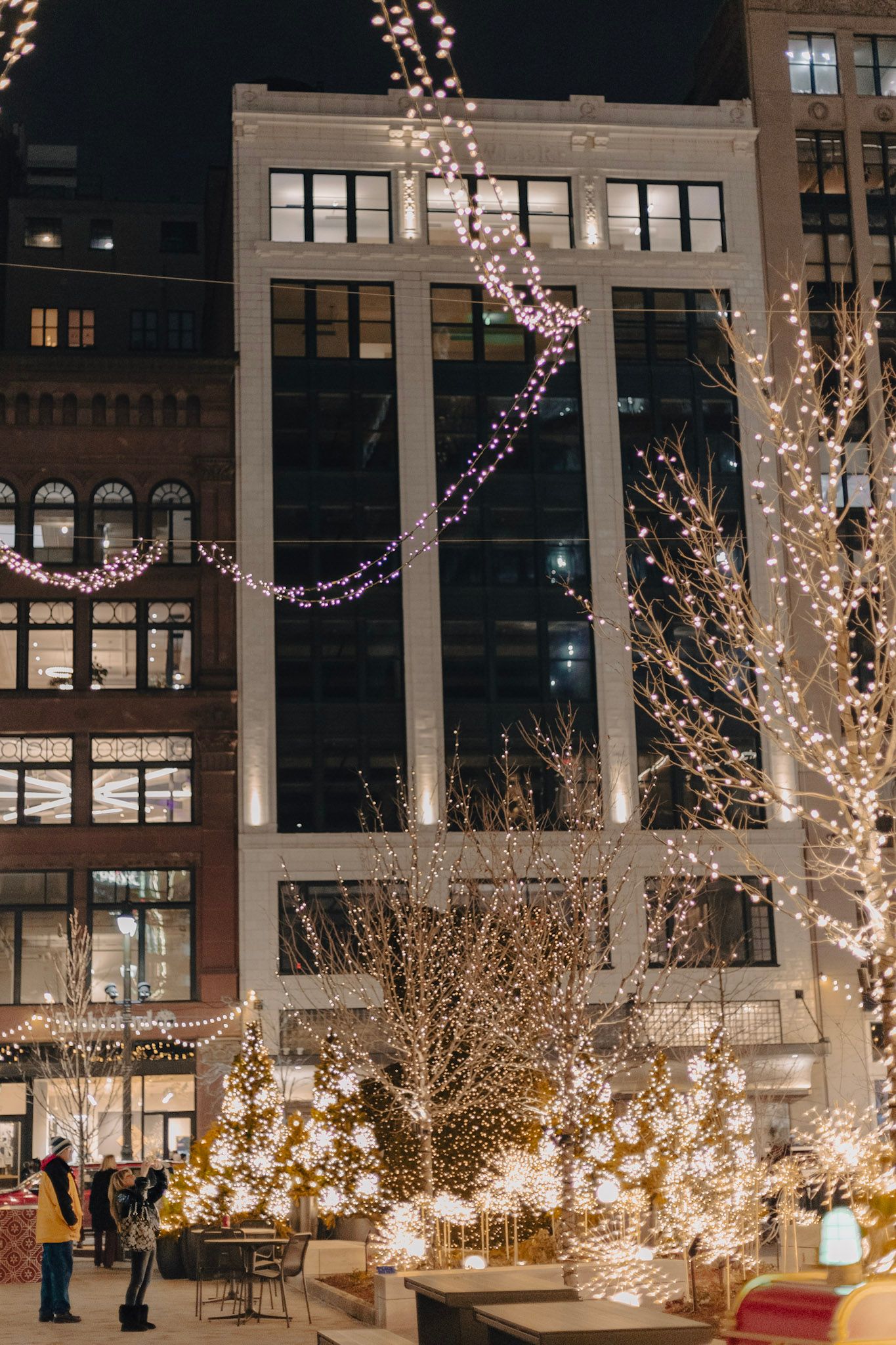 City plaza decorated with holiday lights and snowflake ornaments on trees at night.