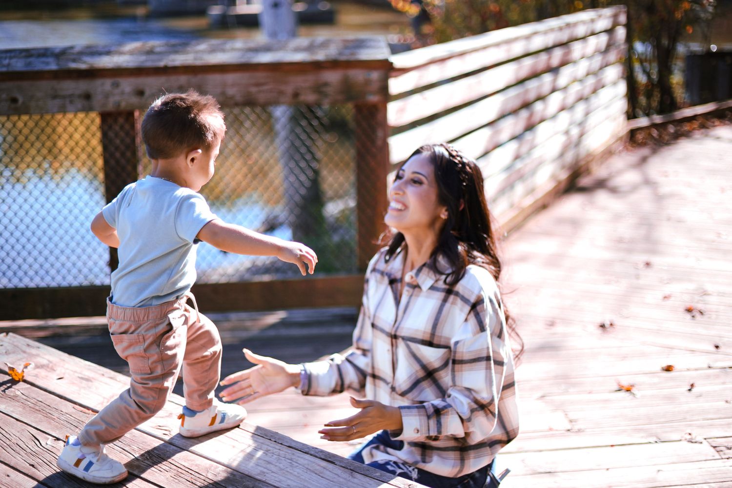 Young mother kneeling on wooden deck reaching out to her toddler son on a sunny autumn day.
