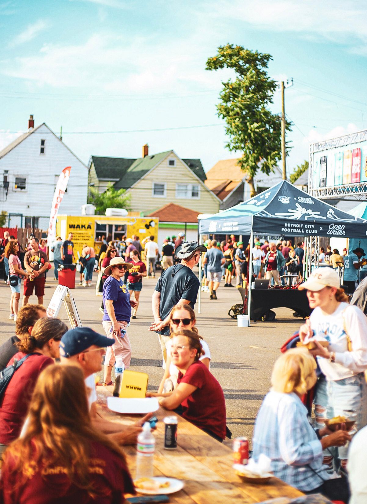 Fans eating and socializing at an outdoor Detroit City FC soccer match street festival on a sunny day.