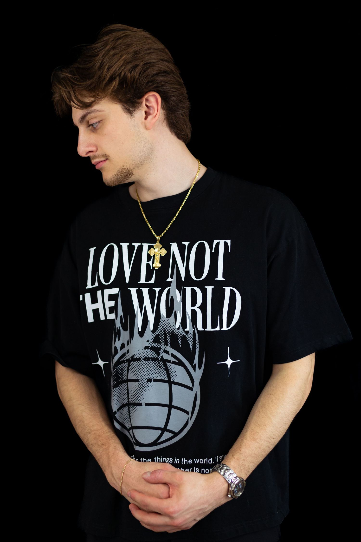 Young man in black Love Not The World graphic tee with gold cross necklace and watch, looking down.