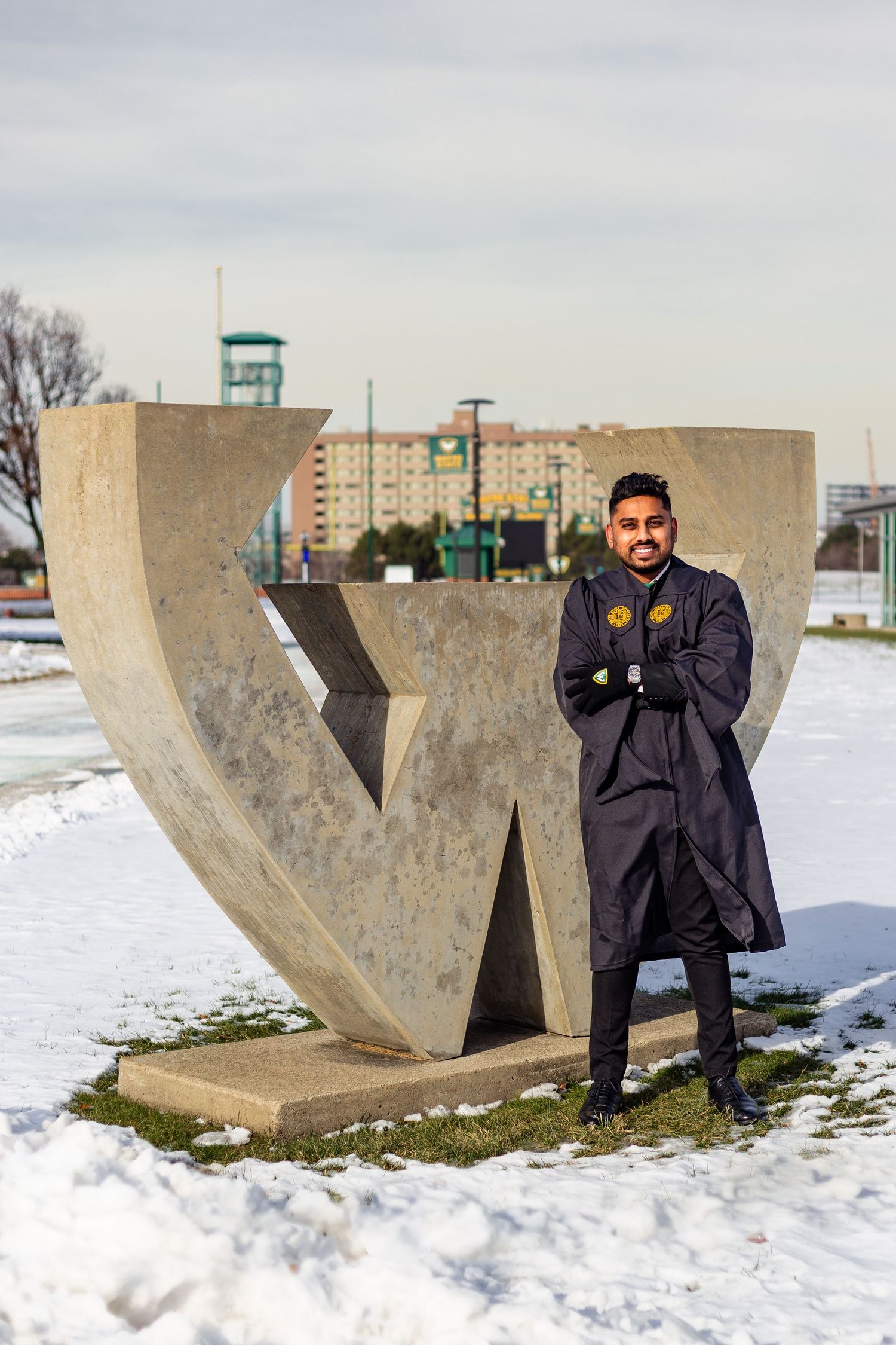 Smiling graduate in black gown posing by W sculpture in snow.