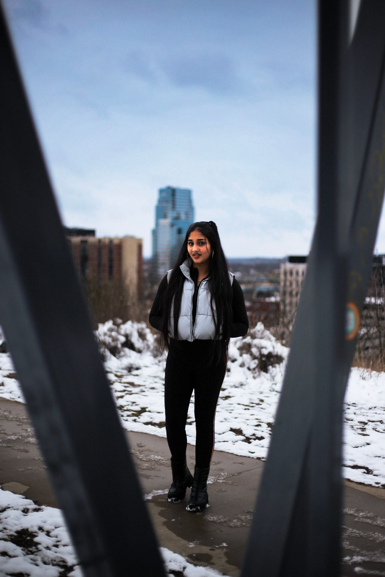 Young woman in gray vest and black pants standing between metal beams with winter city skyline.