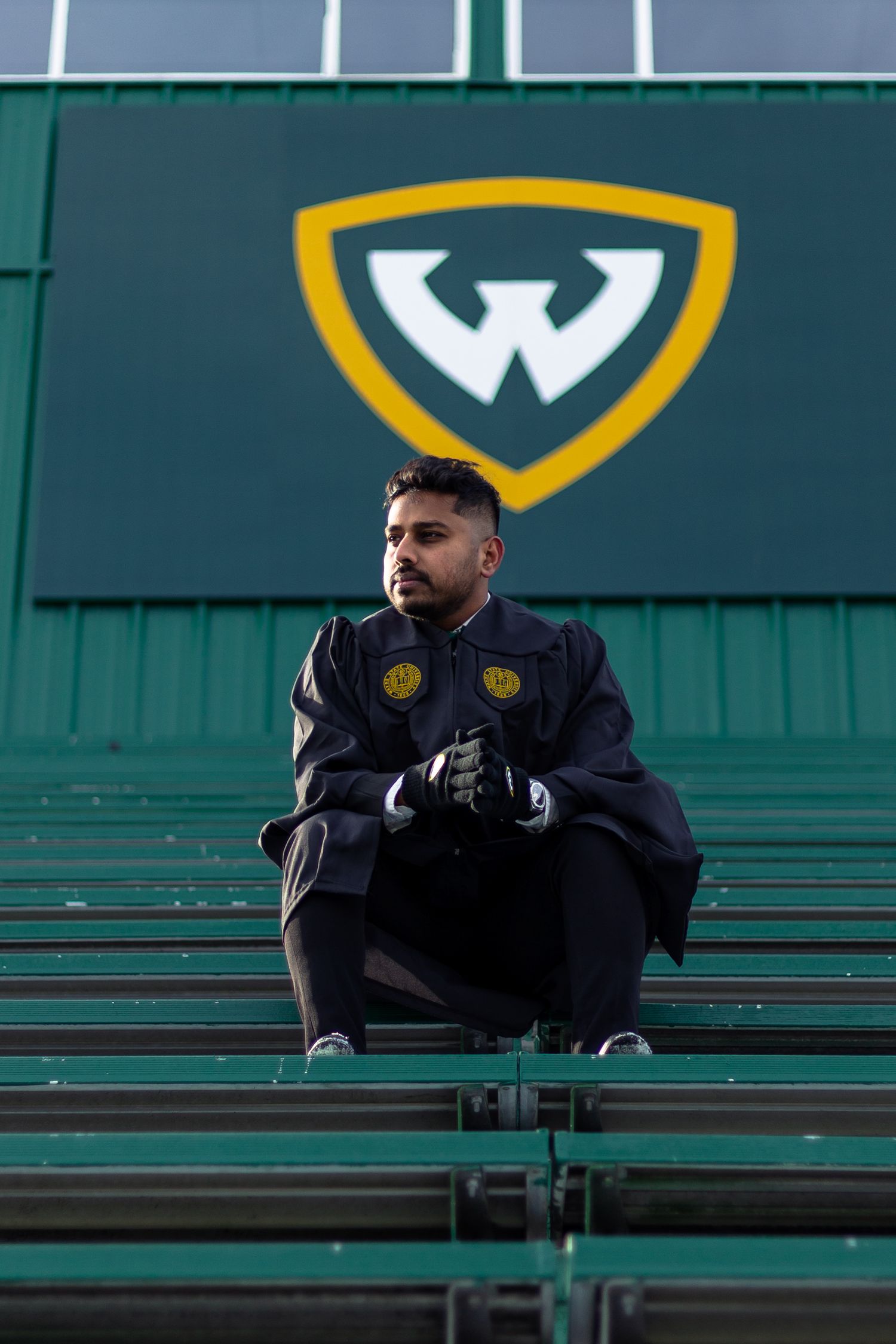 Male graduate in black cap and gown sits in Wayne State University stadium beneath the WSU logo.