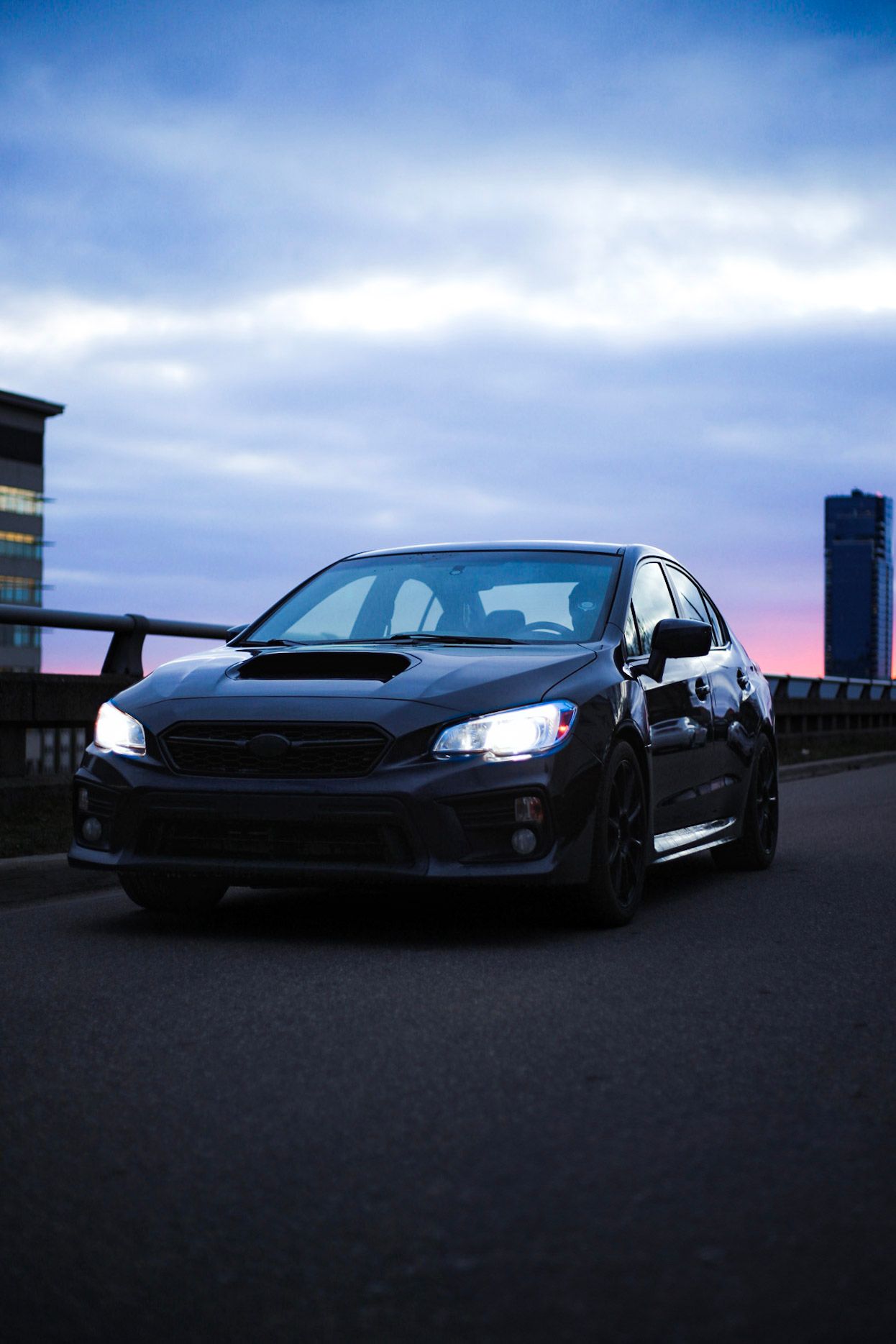 Black Subaru WRX STI driving on road at twilight with city skyline and dramatic blue sky.