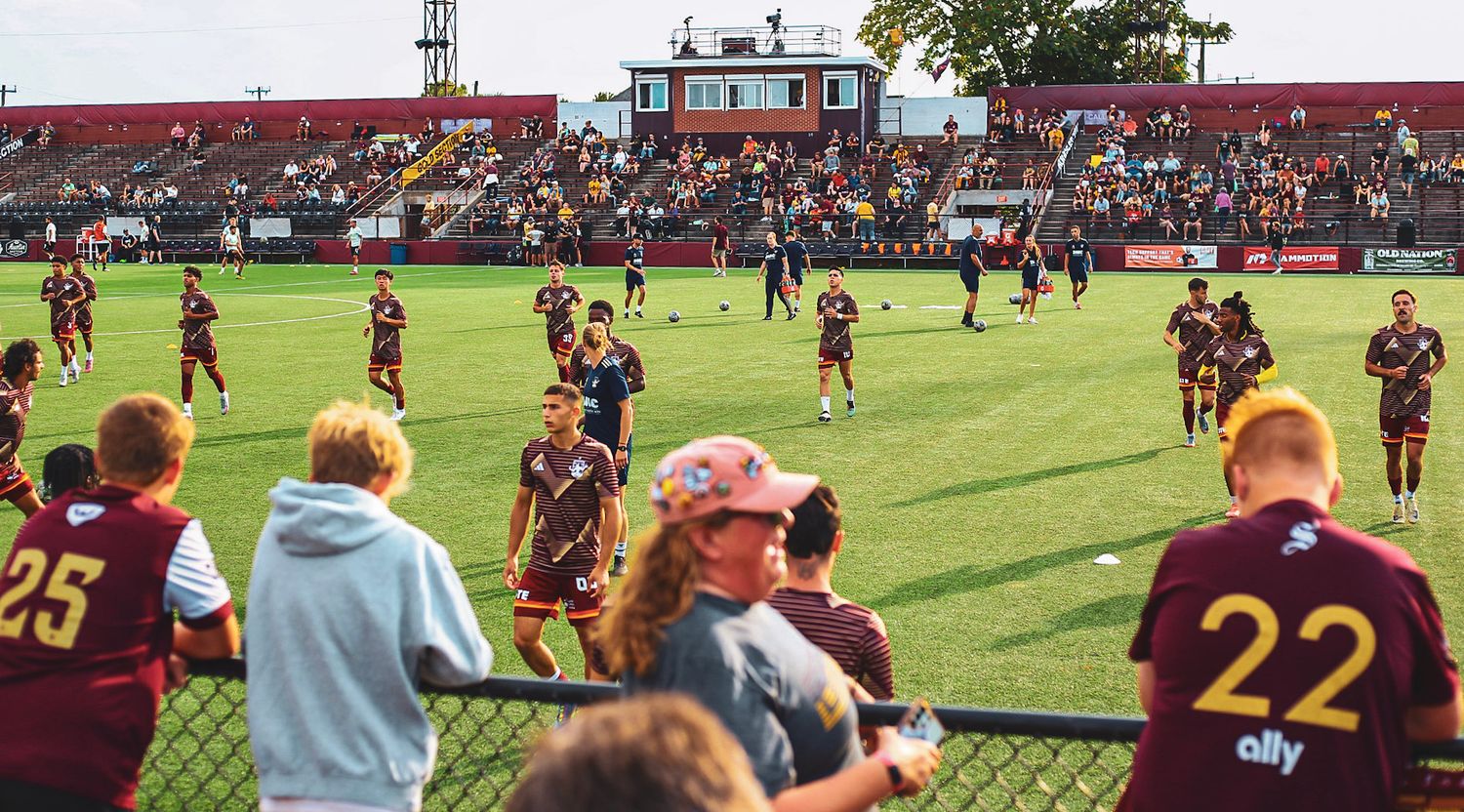 Soccer players in maroon uniforms warming up on a green field while young fans watch from the sideline.