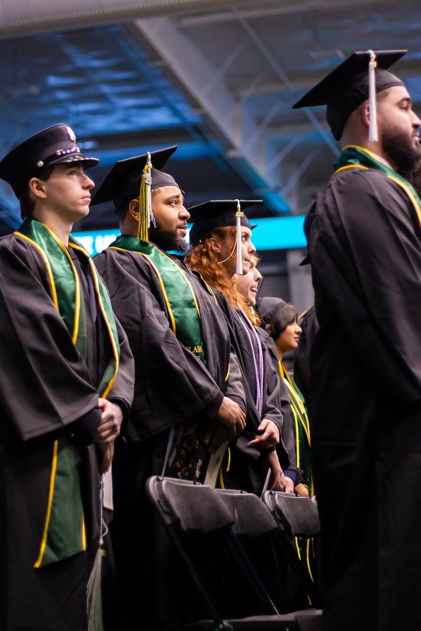 Diverse group of graduates in black robes with green and gold stoles stand at commencement ceremony.
