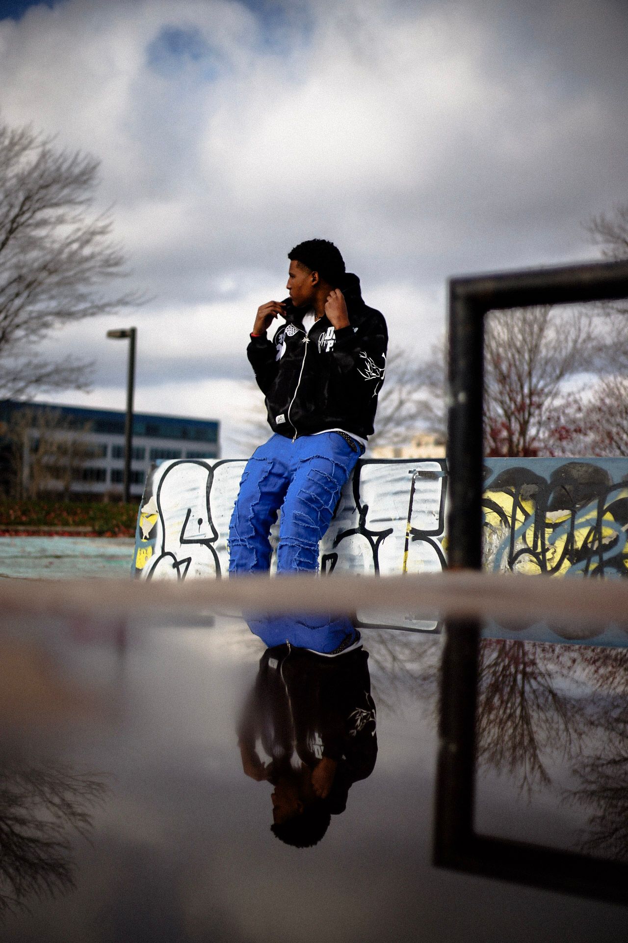 Young man in blue pants reflected in puddle at graffiti-covered skate park under cloudy sky.