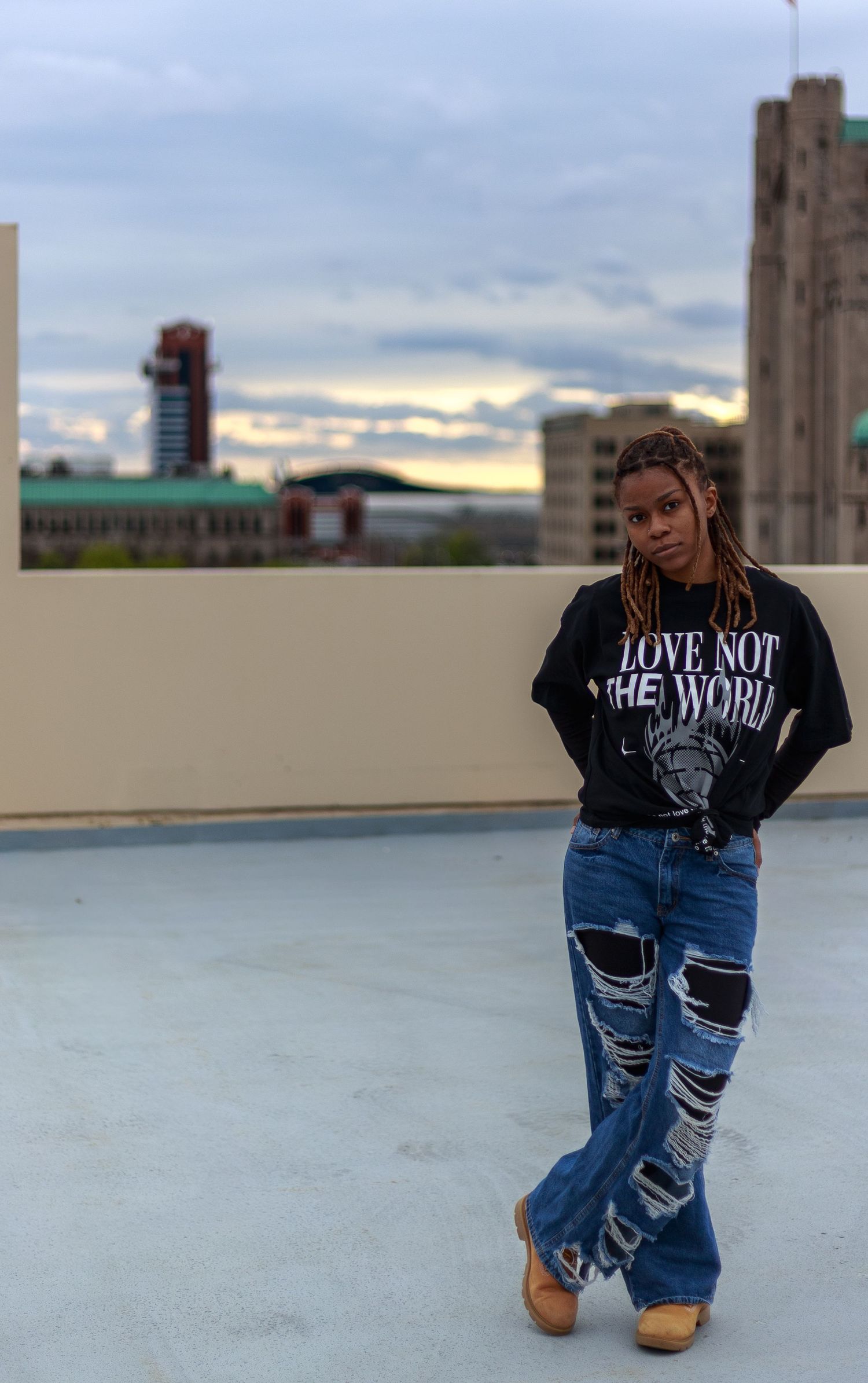 Woman in ripped flare jeans and black graphic tee stands on rooftop with urban skyline at golden hour.