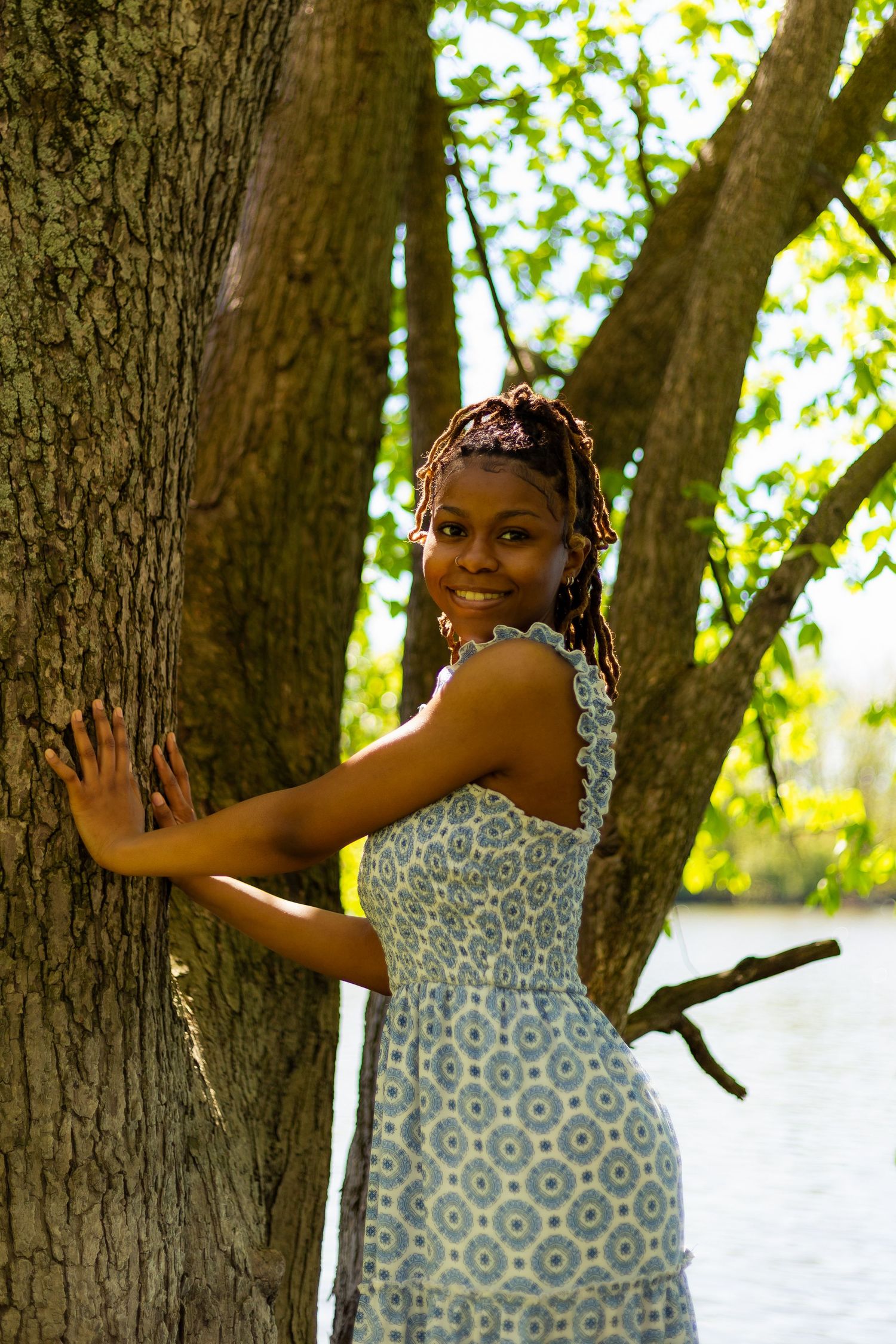 Smiling young Black woman in blue floral dress embraces a large tree trunk in a sunny park by a lake.