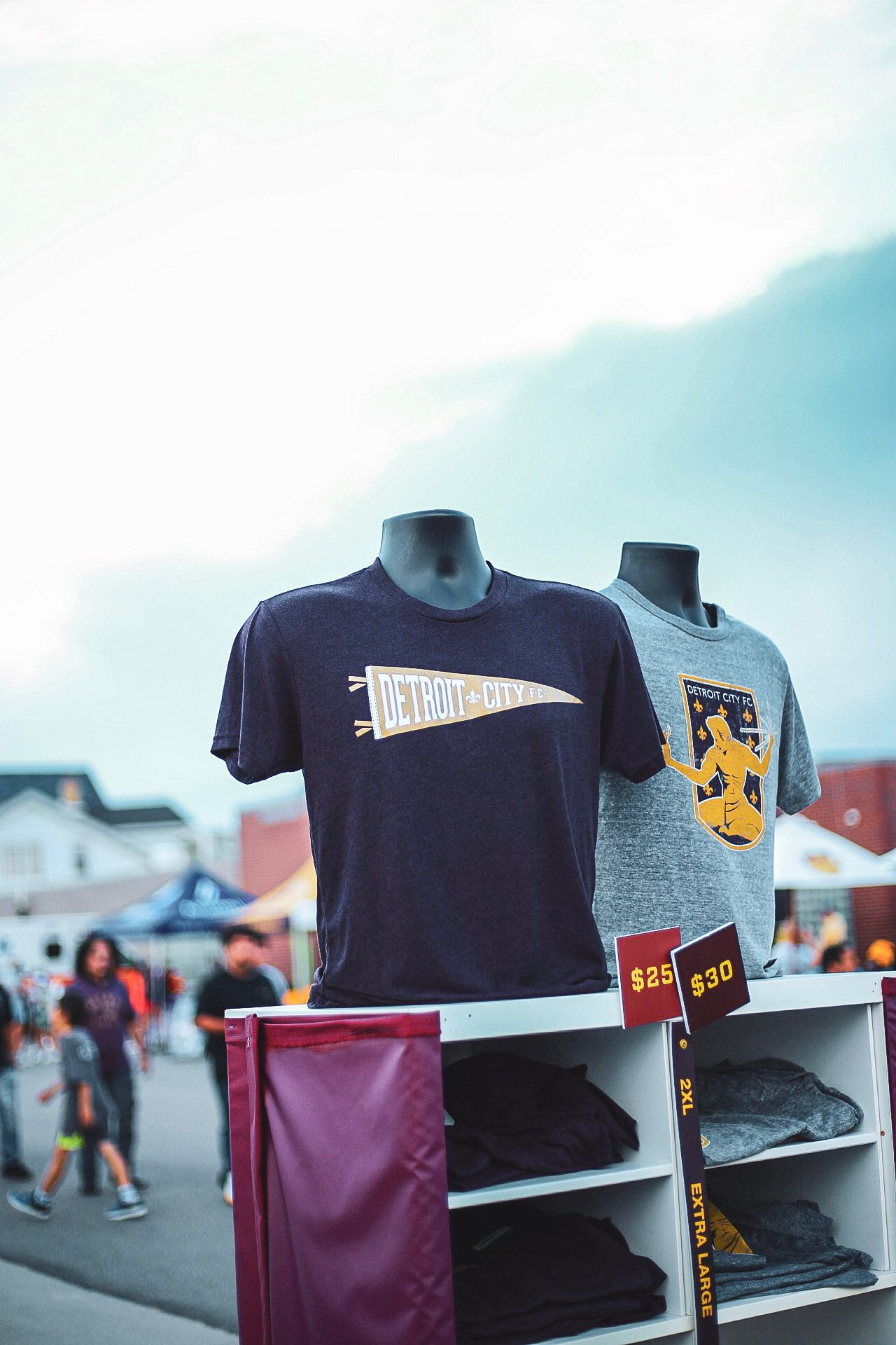 Detroit City FC merchandise display with navy and gray t-shirts priced at $25 and $30 outdoors.