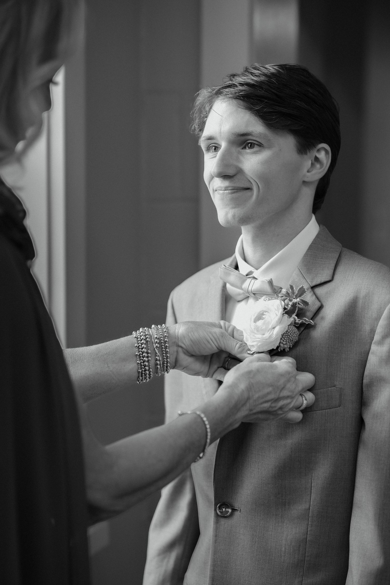Black and white photo of a smiling person in a suit jacket having a floral boutonniere pinned on by another person.