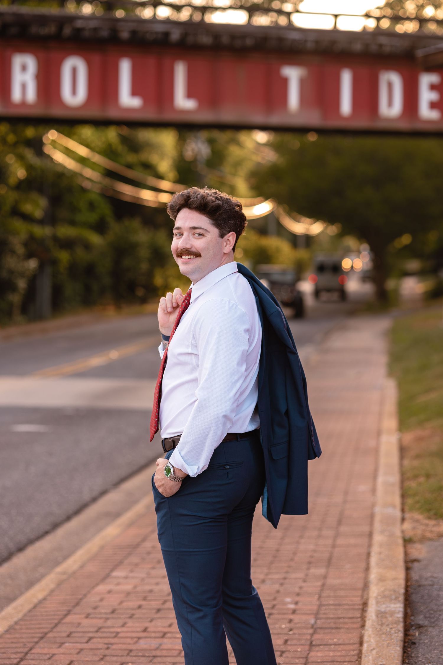 Stylish young man with mustache poses in white shirt and red tie, holding navy blazer, near Roll Tide bridge.