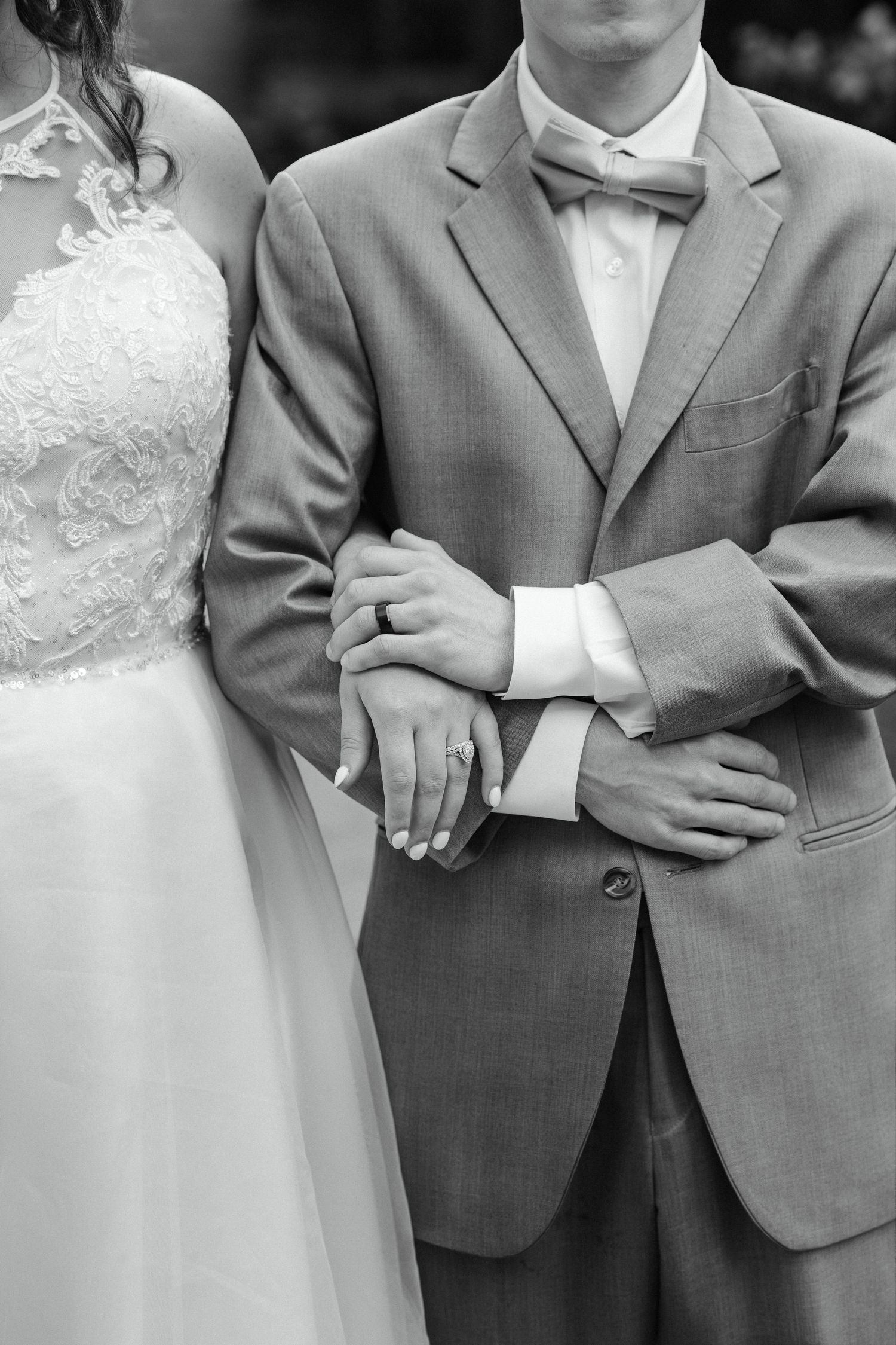 Black and white photo of bride and groom standing together, showing their clasped hands and wedding rings.