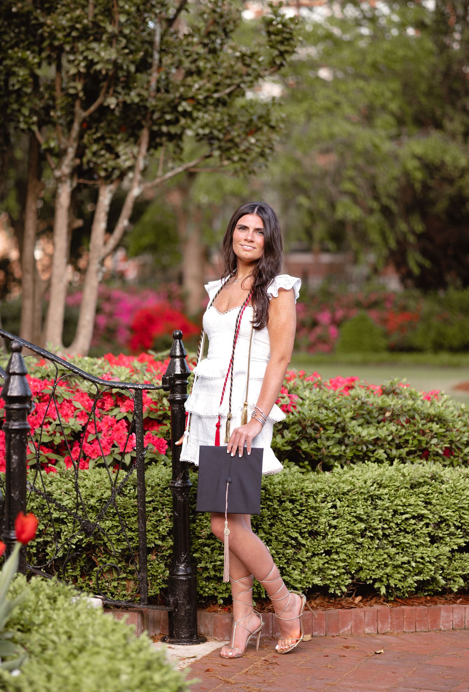 Graduate in white dress poses by garden path with red flowers and iron fencing.